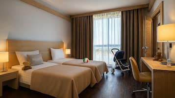 Ein Hotelzimmer mit zwei Betten, einem Schreibtisch, einem Stuhl, einer Lampe, einem Teddybären, einem Kinderwagen und einem Fenster mit Vorhängen.