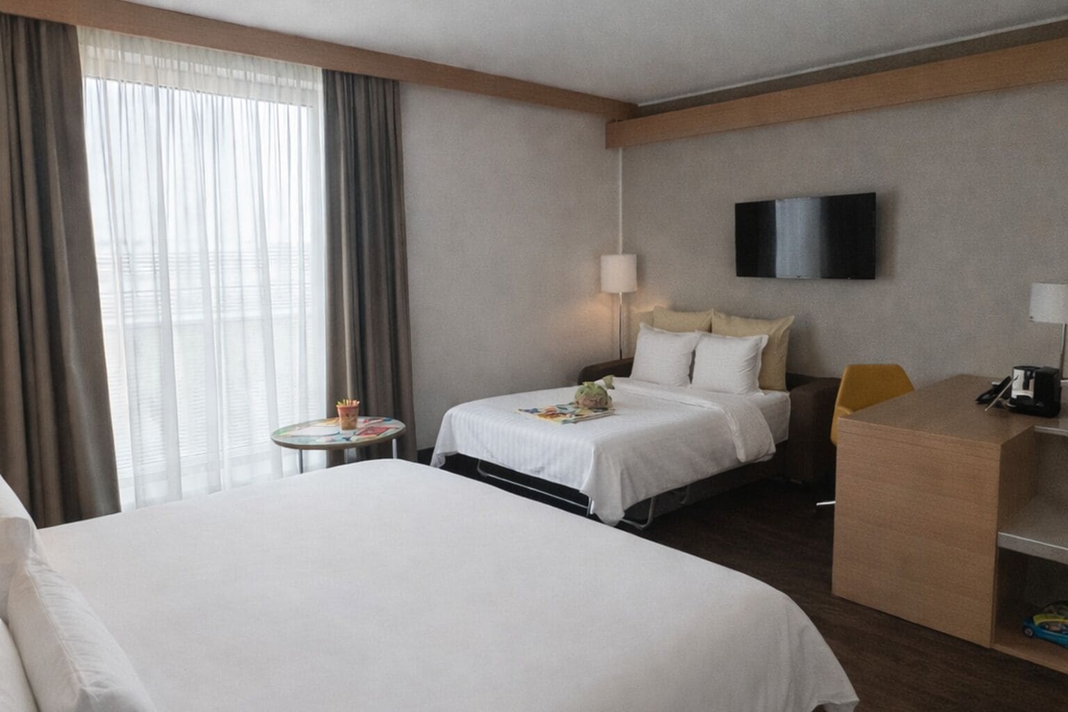 Habitación de hotel con una cama grande, una cama más pequeña, un escritorio, una silla, un televisor montado en la pared y una ventana con cortinas.