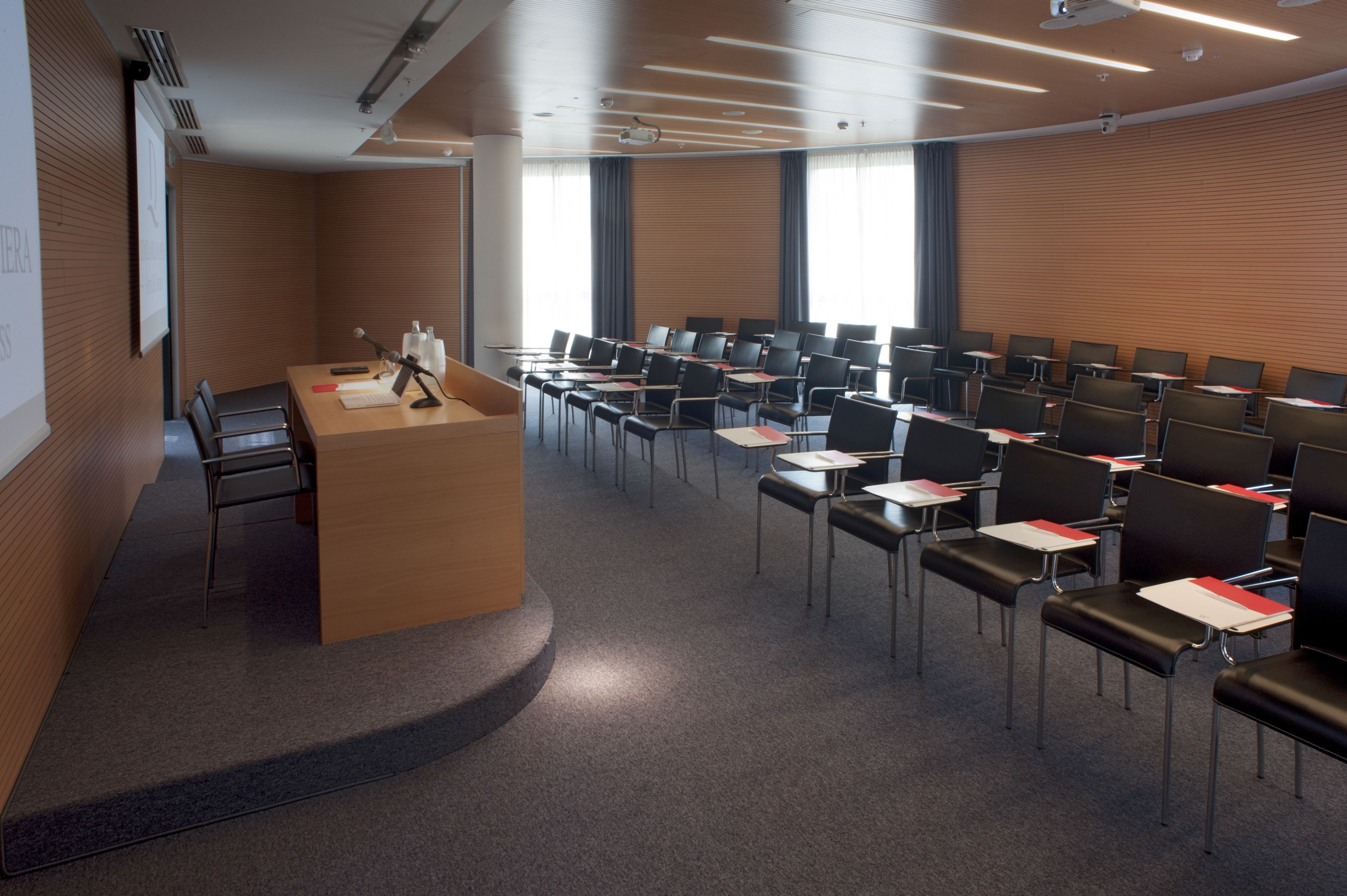 Un salón de conferencias con un atril, filas de sillas y una pantalla grande.