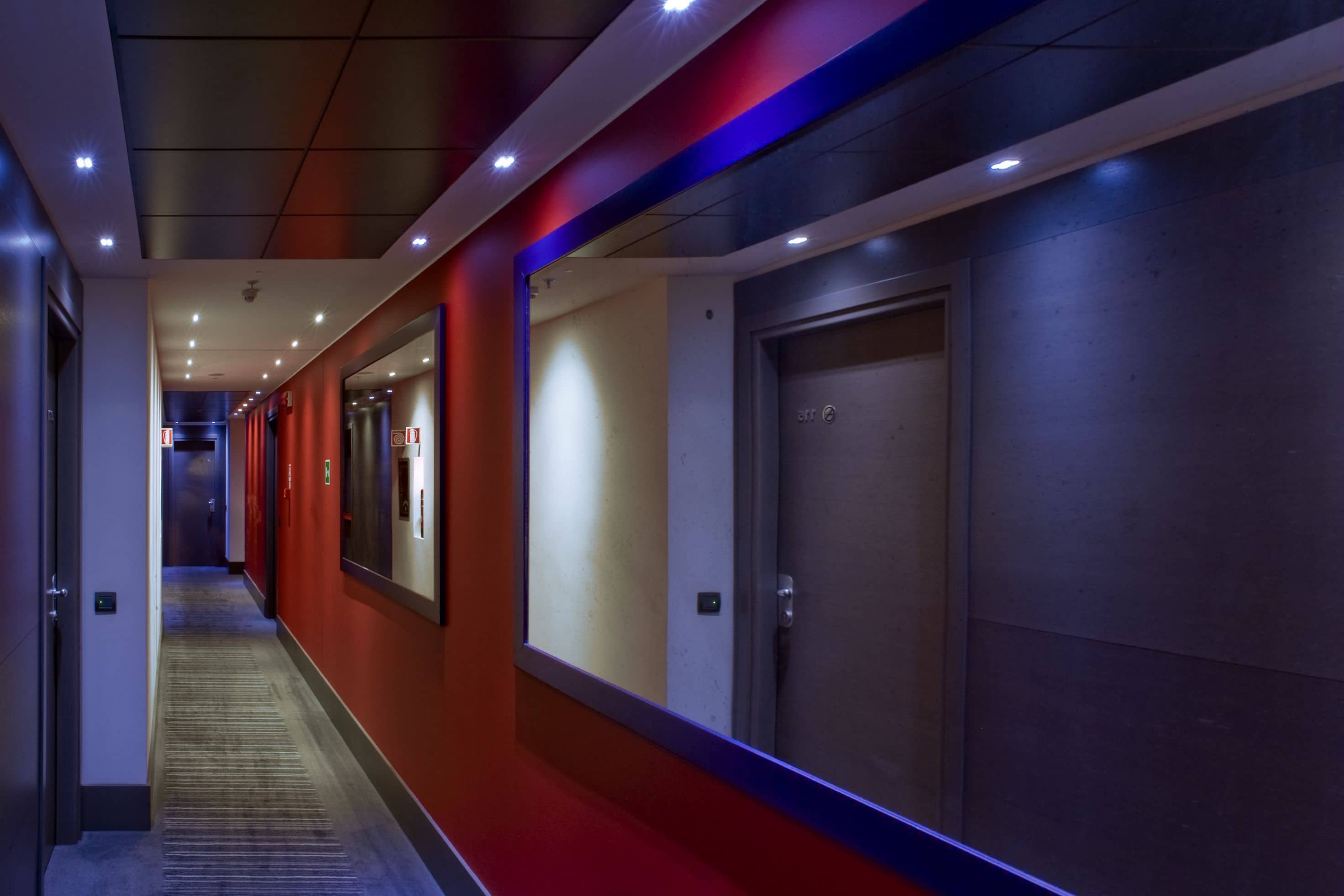 A modern hotel corridor with a red carpet, a mirrored wall, and a door with a sign.