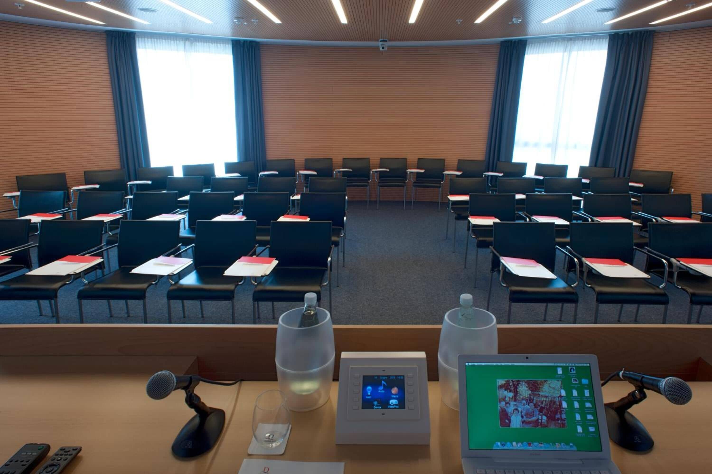 Une salle de conférence avec des rangées de chaises, un pupitre équipé de microphones, un ordinateur portable et un thermostat.