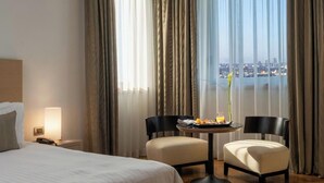 A hotel room with a bed, a bedside table with a lamp, a small table with a chair, and a view of a cityscape through large windows. - Domina Milano Fiera (Novate Milanese)