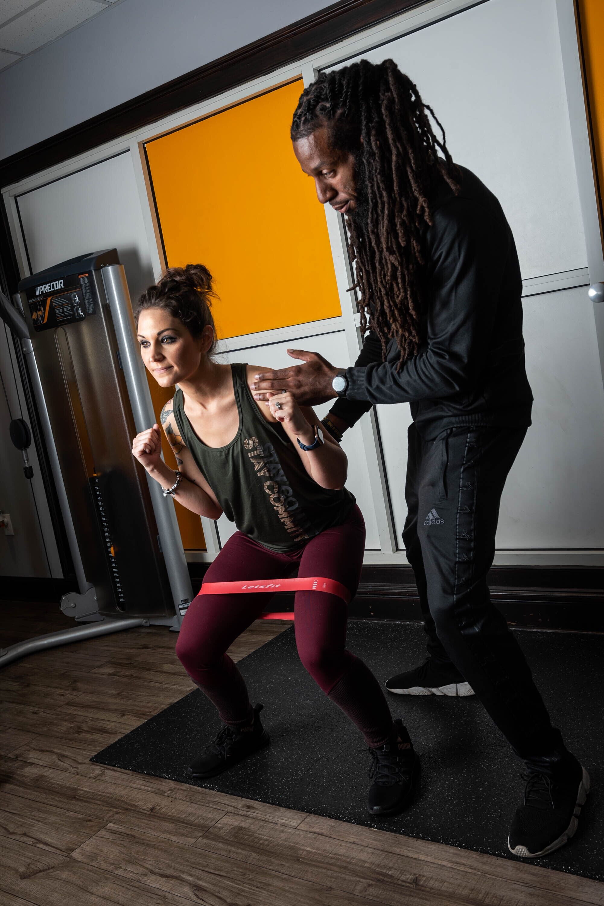 Een sportschool met trainingsapparatuur, een vrouw die squats doet en een man die haar helpt.