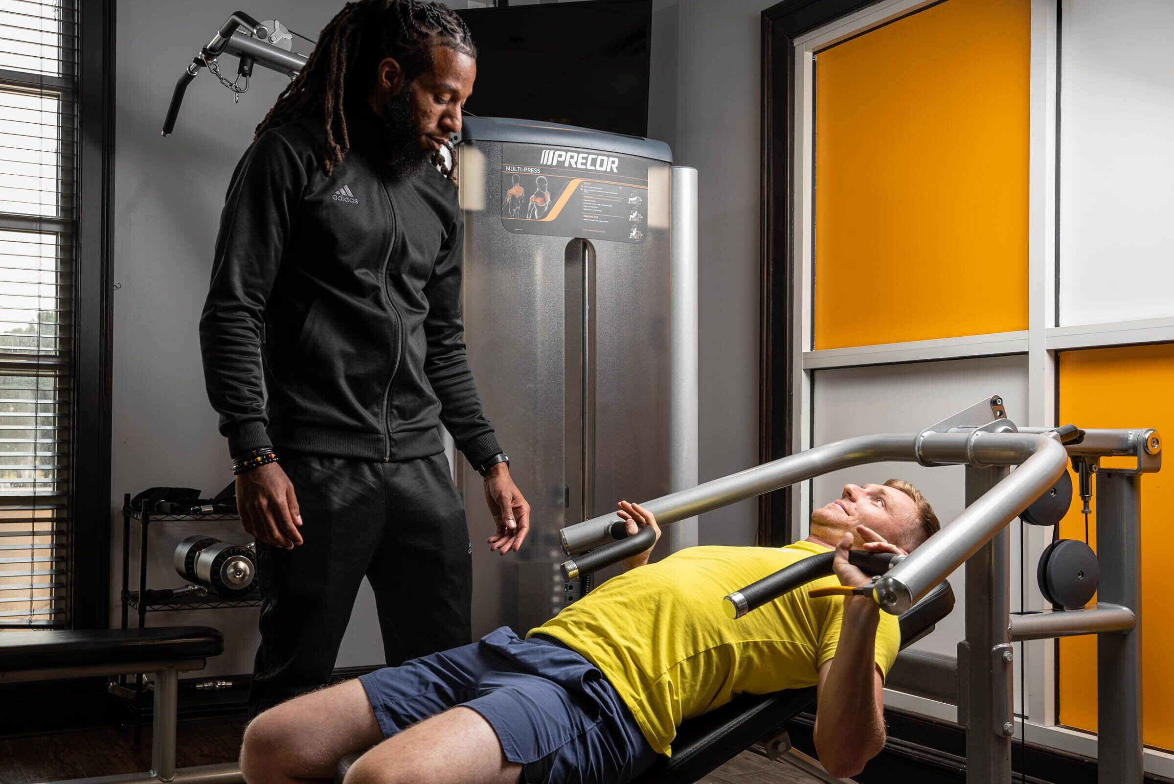A gym with a person lying on a bench, another person standing beside them, and a large exercise machine.