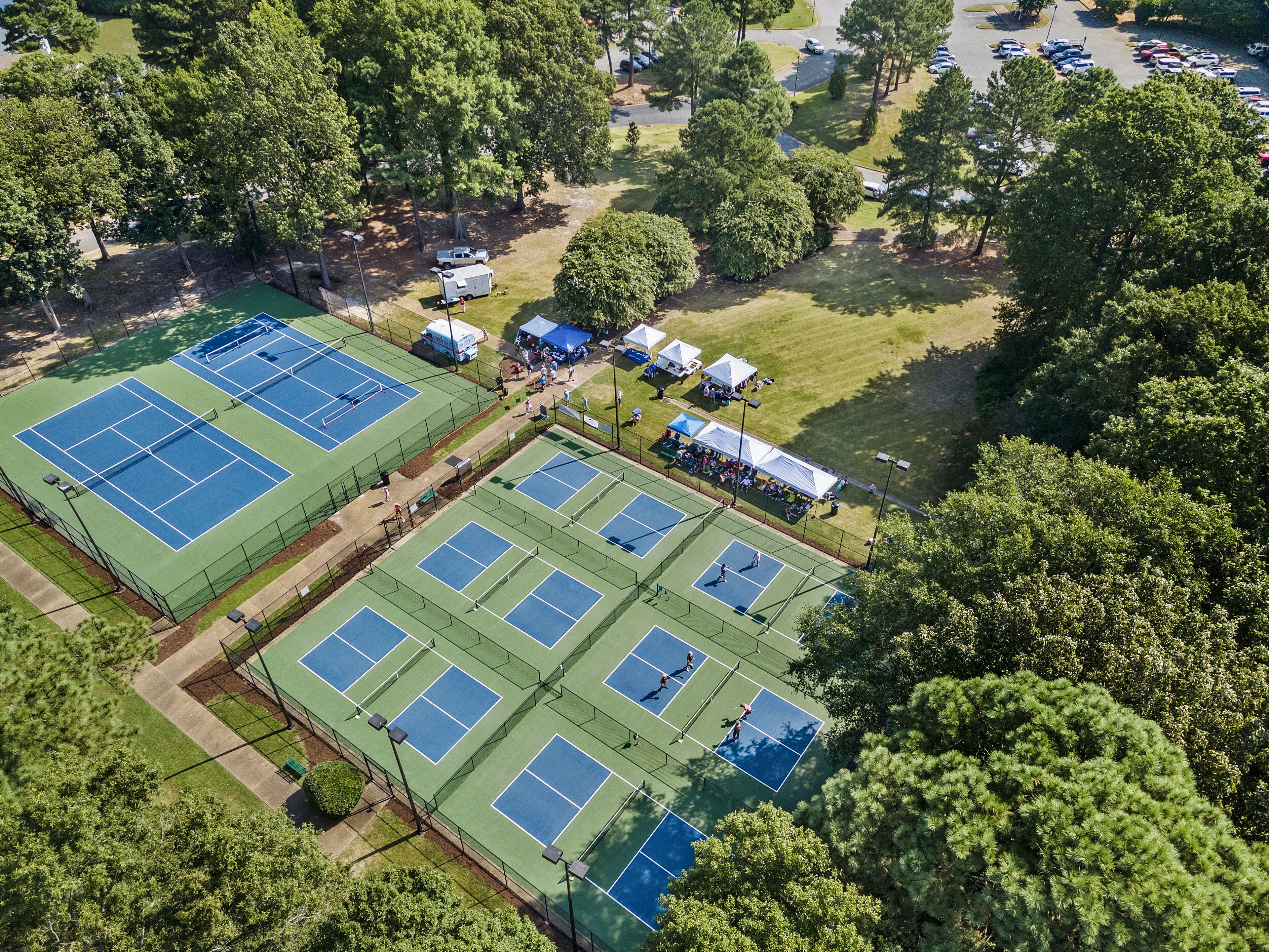 An aerial view of a tennis court complex with multiple courts, surrounded by trees and a parking area.