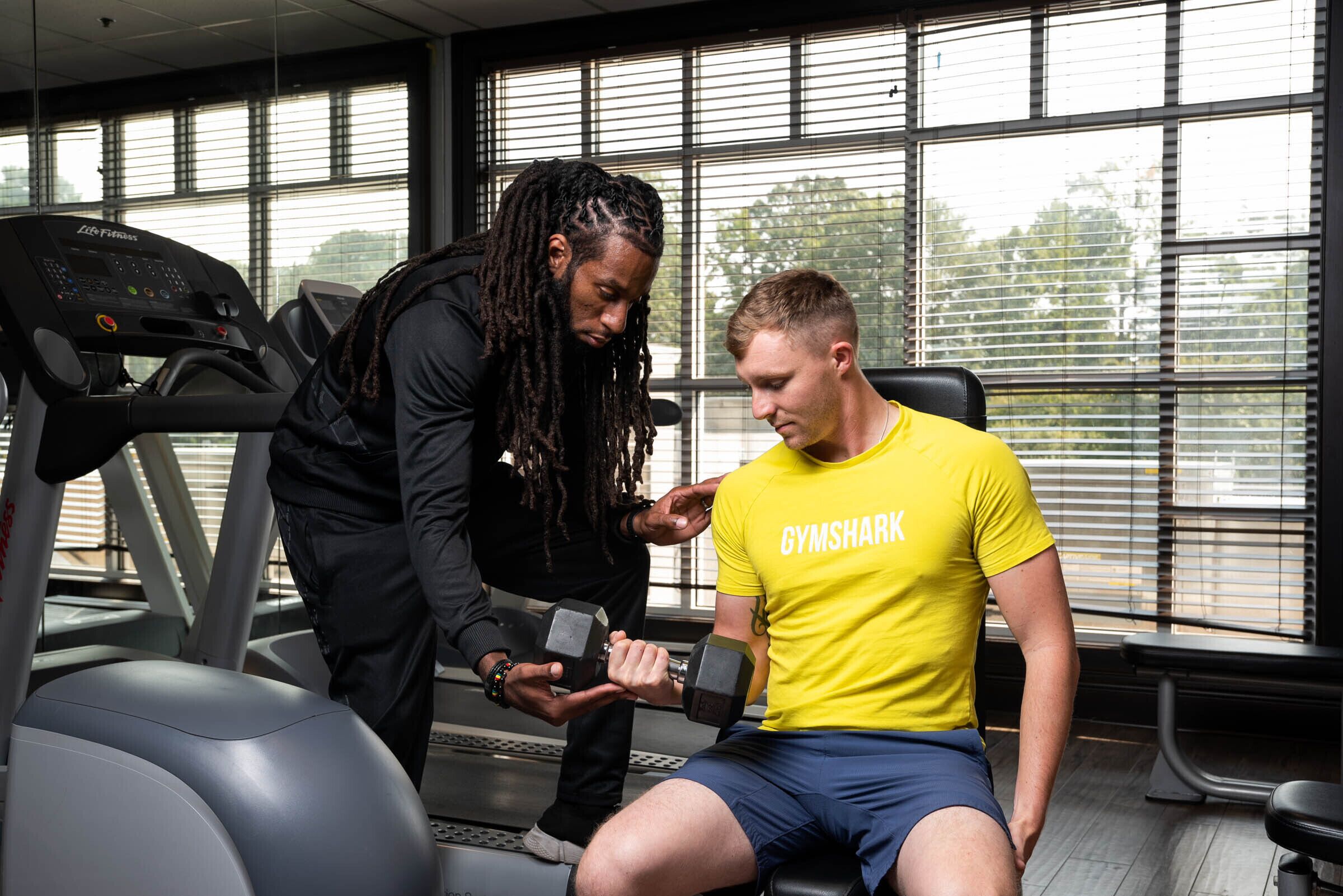 A gym with a treadmill, a seated individual, and a standing person assisting with an exercise.