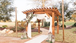 A wooden pergola with a white draped fabric and floral decorations, set in a landscaped garden with a gravel path leading to it. - The Lodge at Kiln Creek Resort (Newport News)