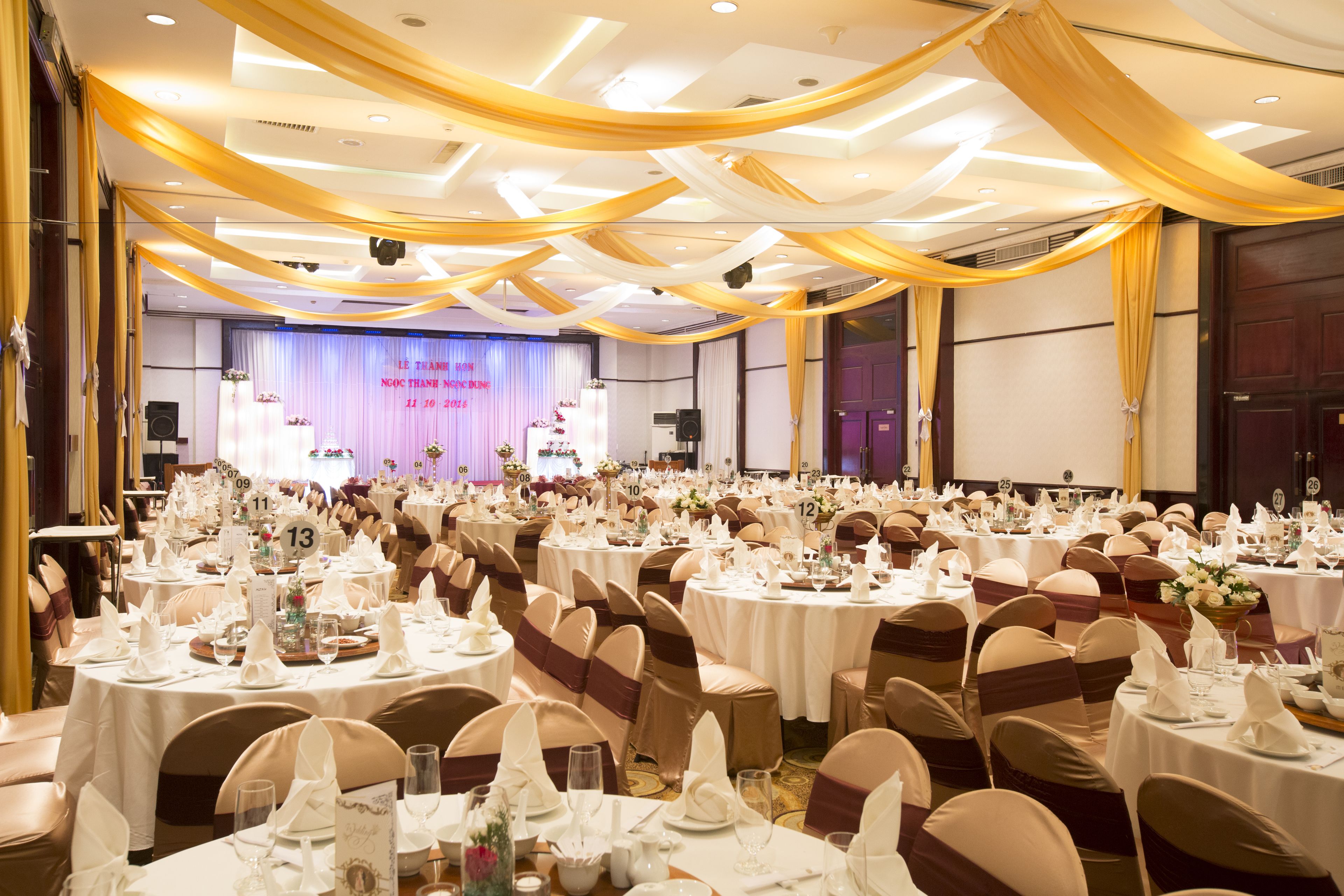 Une salle de banquet avec des tables rondes dressées pour un événement formel, agrémentée de nappes blanches et de tentures dorées.