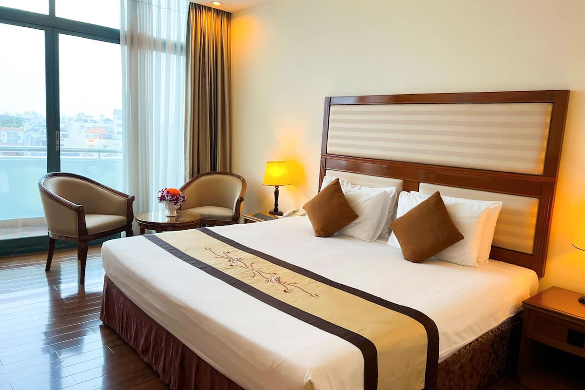 Une chambre d’hôtel avec un grand lit, deux fauteuils, une table de chevet avec une lampe, et une vue sur la ville grâce à de grandes fenêtres.