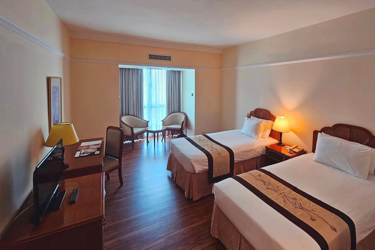 Une chambre d’hôtel avec deux lits, un bureau avec une lampe, une chaise et une fenêtre avec des rideaux.