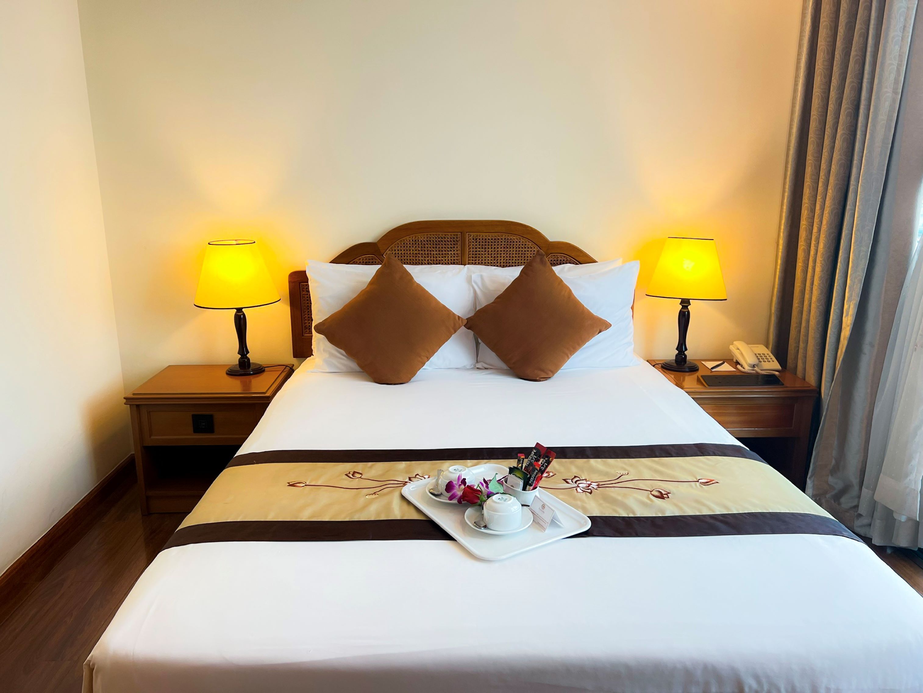 A neatly made hotel bed with two pillows, a tray with a teapot and flowers, and two bedside lamps.