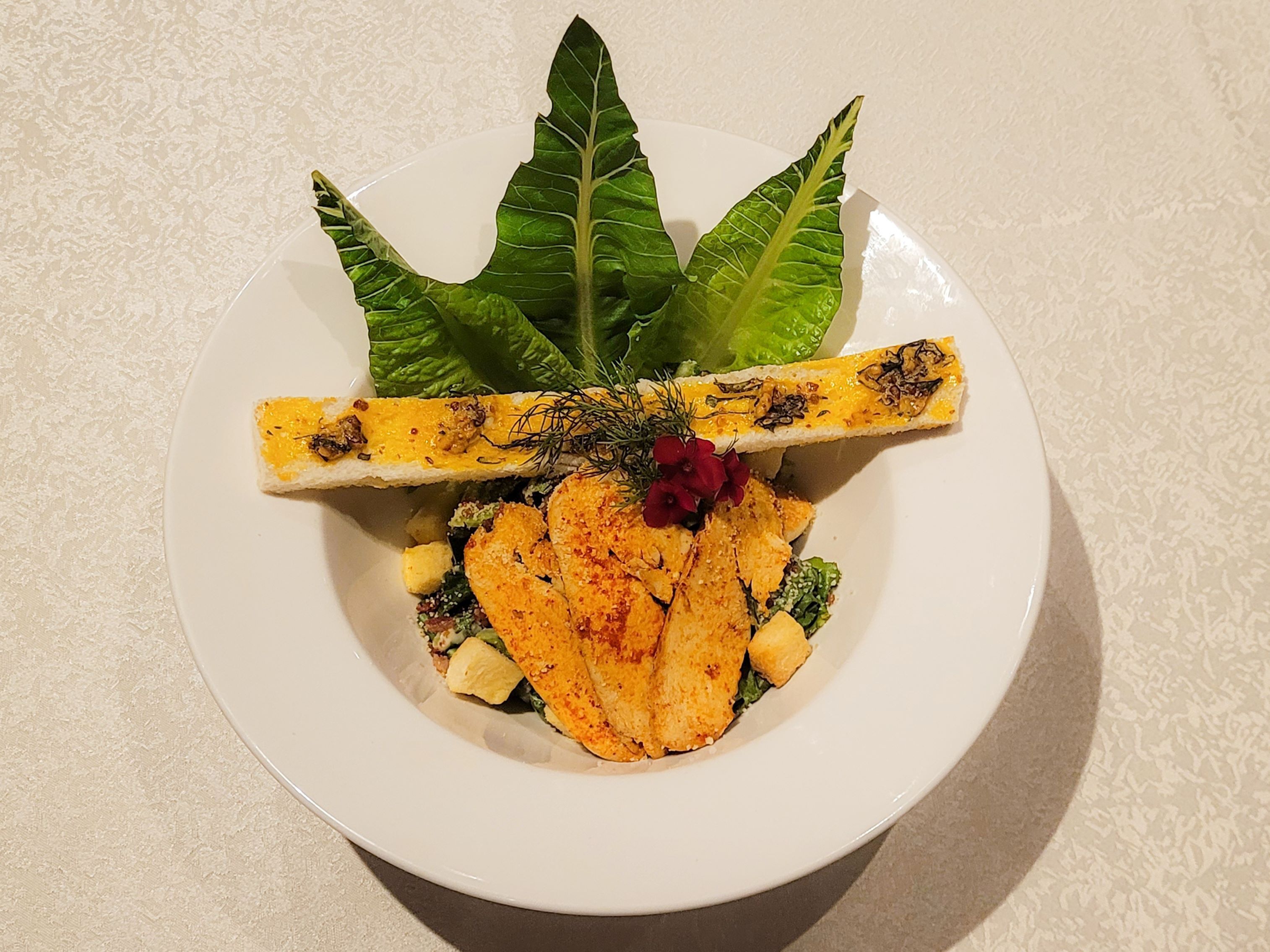 A plate with grilled chicken, vegetables, and a piece of grilled bread, garnished with herbs and flowers.