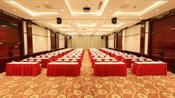 Une salle de conférence avec des rangées de tables, chacune recouverte d’une nappe rouge et agrémentée de bouteilles d’eau. La salle est dotée d’un tapis à motifs et d’un plafond moderne avec des luminaires encastrés.