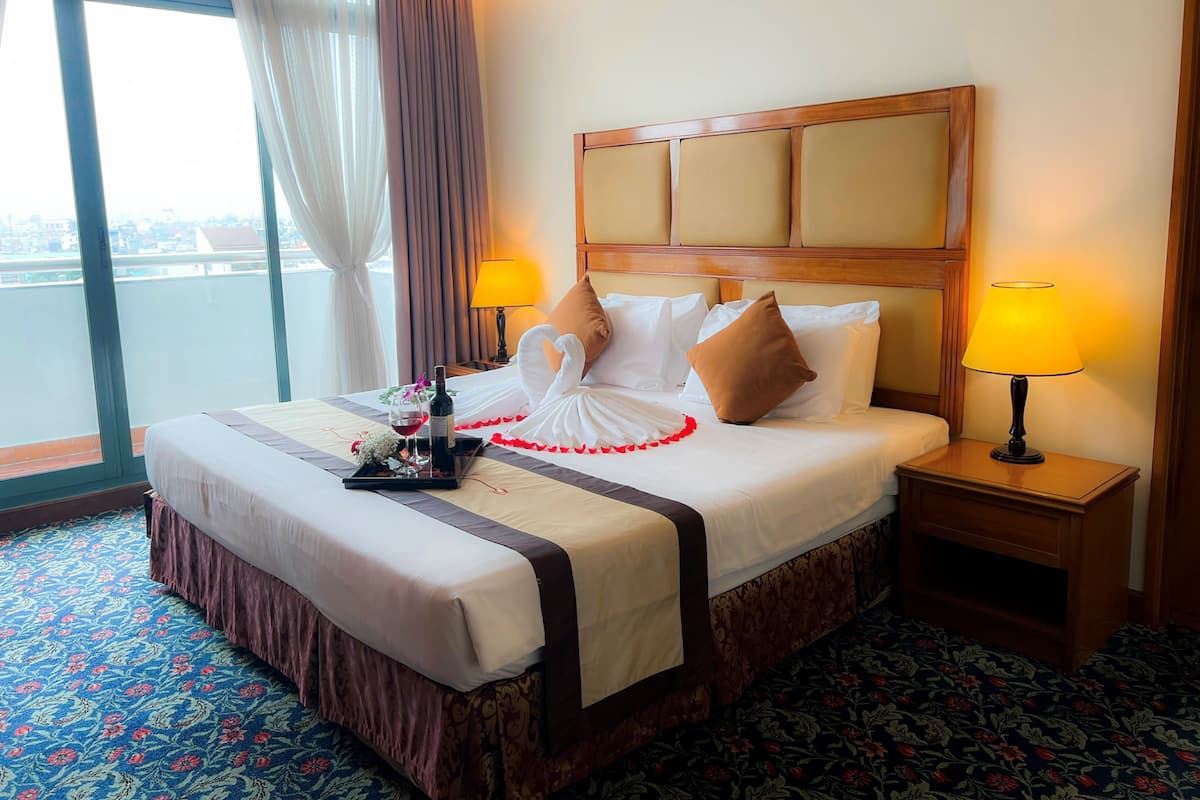 Une chambre d’hôtel avec un grand lit, deux lampes de chevet, un plateau avec une bouteille et un verre, et une vue dégagée sur l’extérieur grâce à une grande fenêtre.
