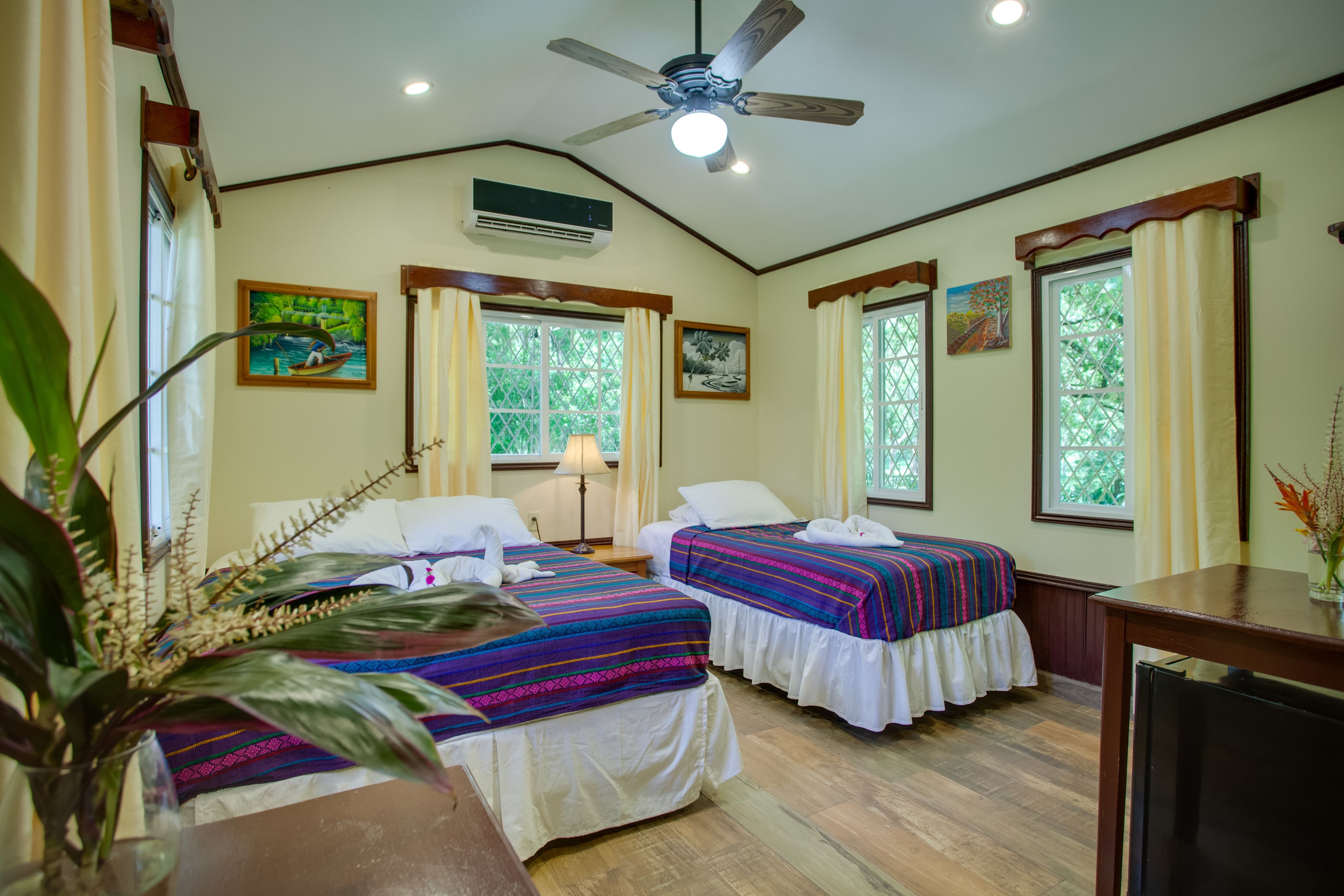 A room with two beds, a ceiling fan, an air conditioner, and large windows with curtains.