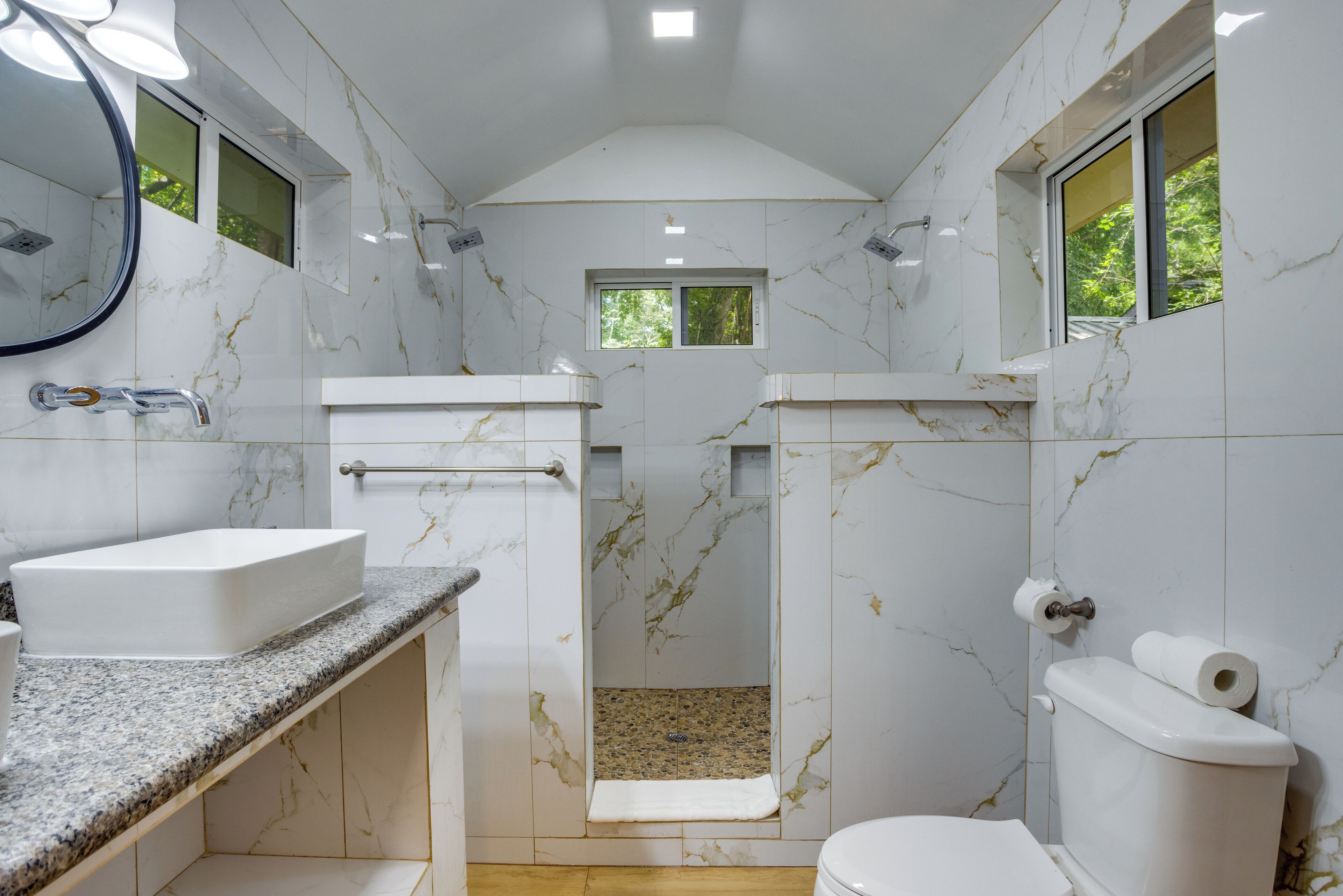 A bathroom with a white sink, toilet, and shower. There are two windows providing natural light.