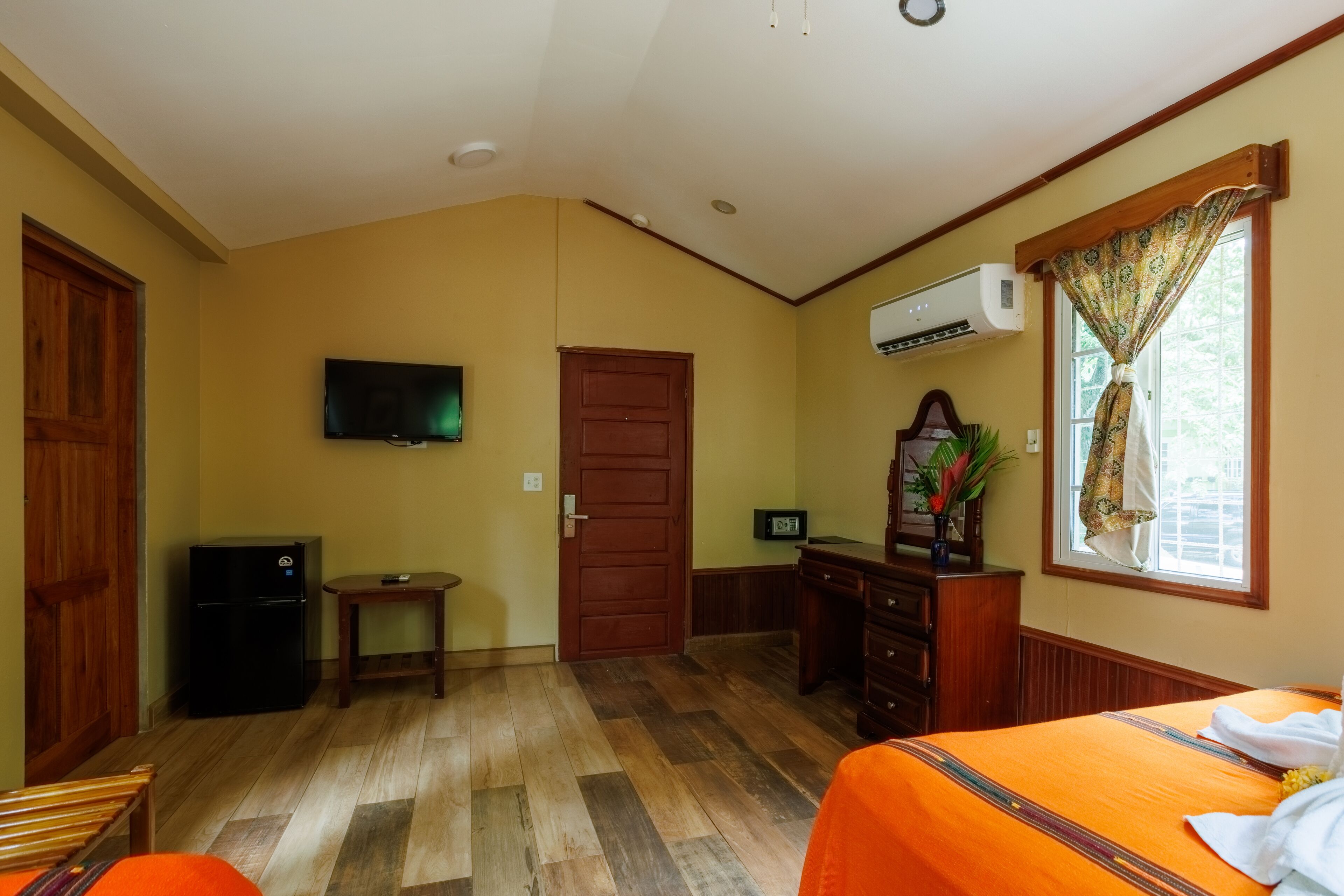 Ein Hotelzimmer mit Holzboden, einem Bett mit orangefarbener Bettdecke, einem Schreibtisch mit Computer, einem an der Wand befestigten Fernseher und einem Fenster mit Vorhängen.