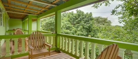 Un espace extérieur aménagé, abrité et doté de chaises et de tables en bois, entouré d’une rambarde verte et offrant une vue sur une végétation luxuriante.