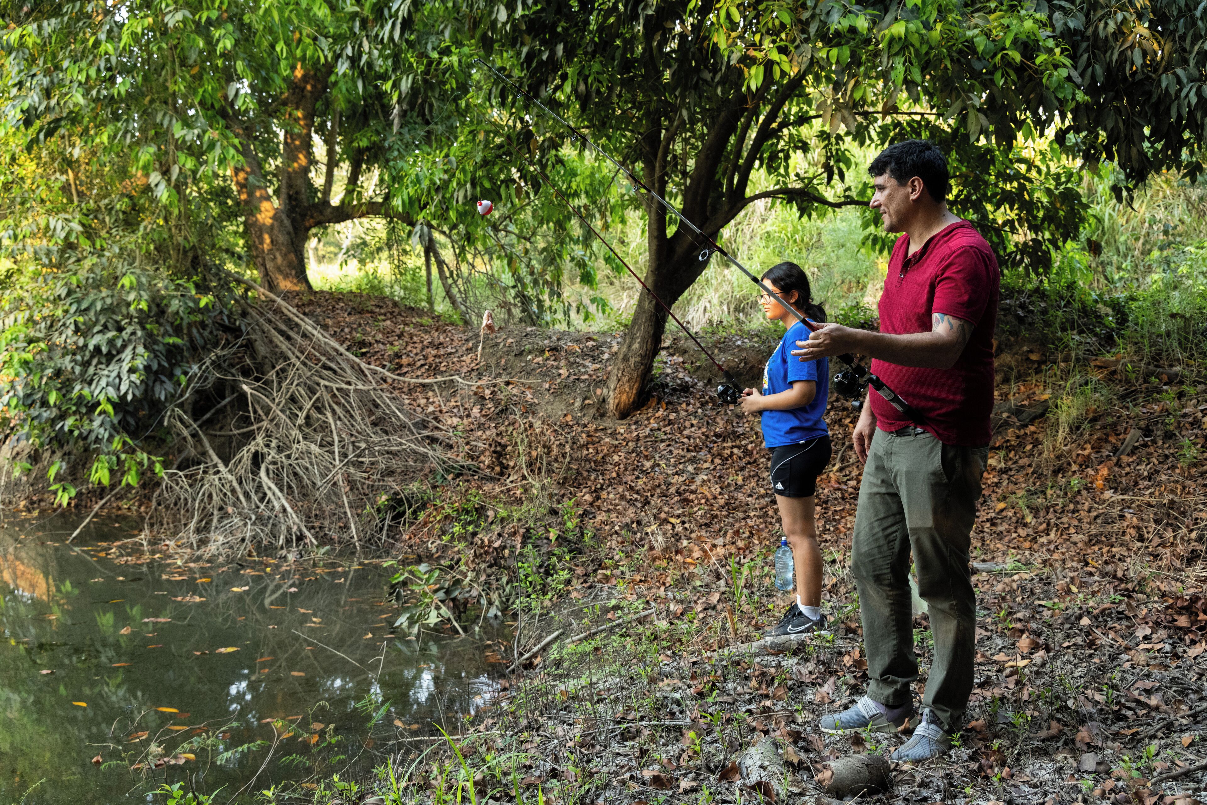 Two people fishing by a calm river surrounded by trees.