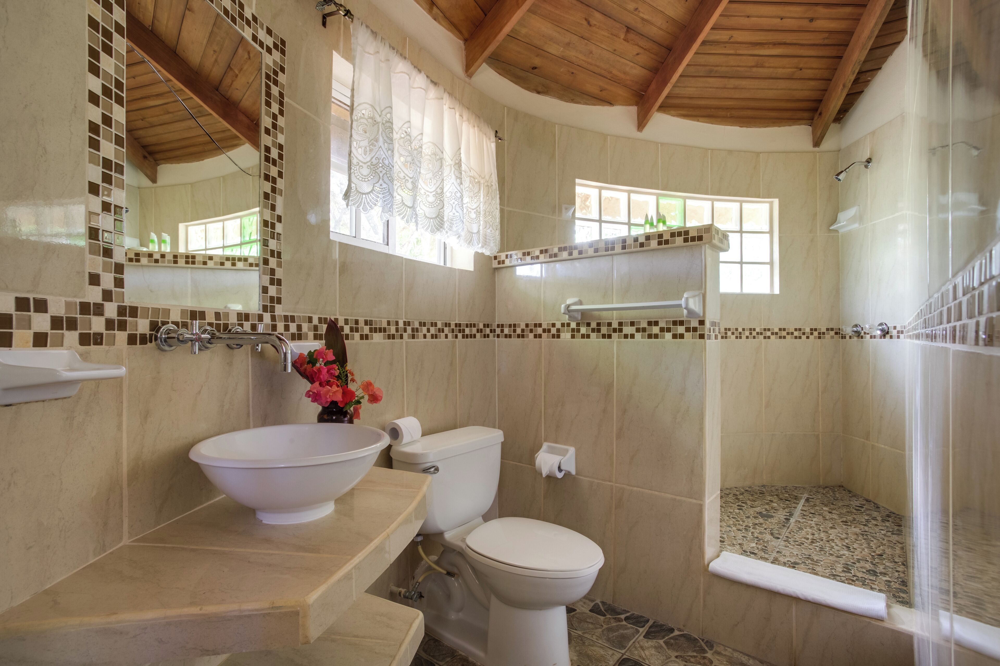 A bathroom with a white sink, toilet, and shower. It features tiled walls, a wooden ceiling, and a window.