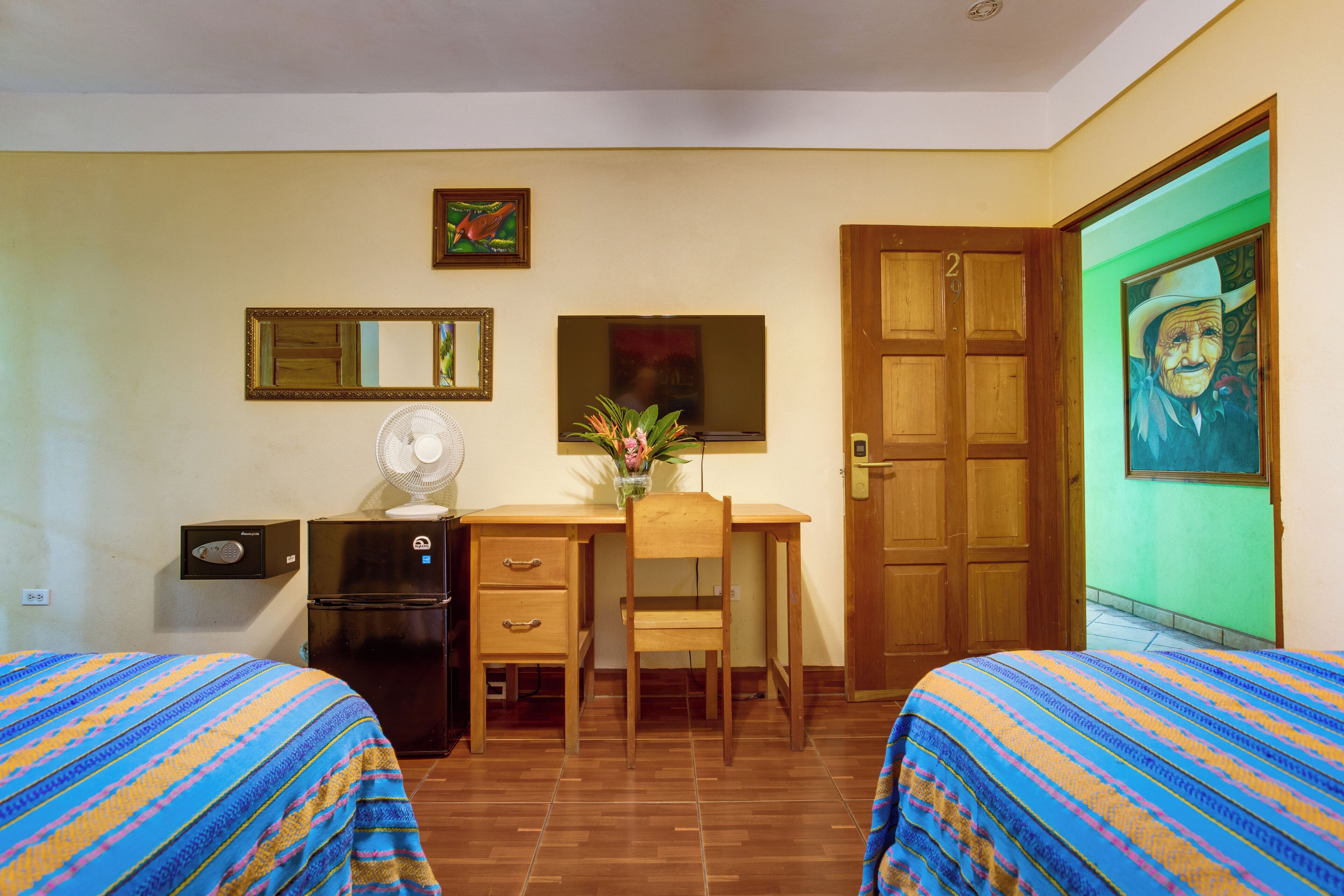 Una habitación con dos camas, un escritorio, una silla, un ventilador, un refrigerador y una puerta.