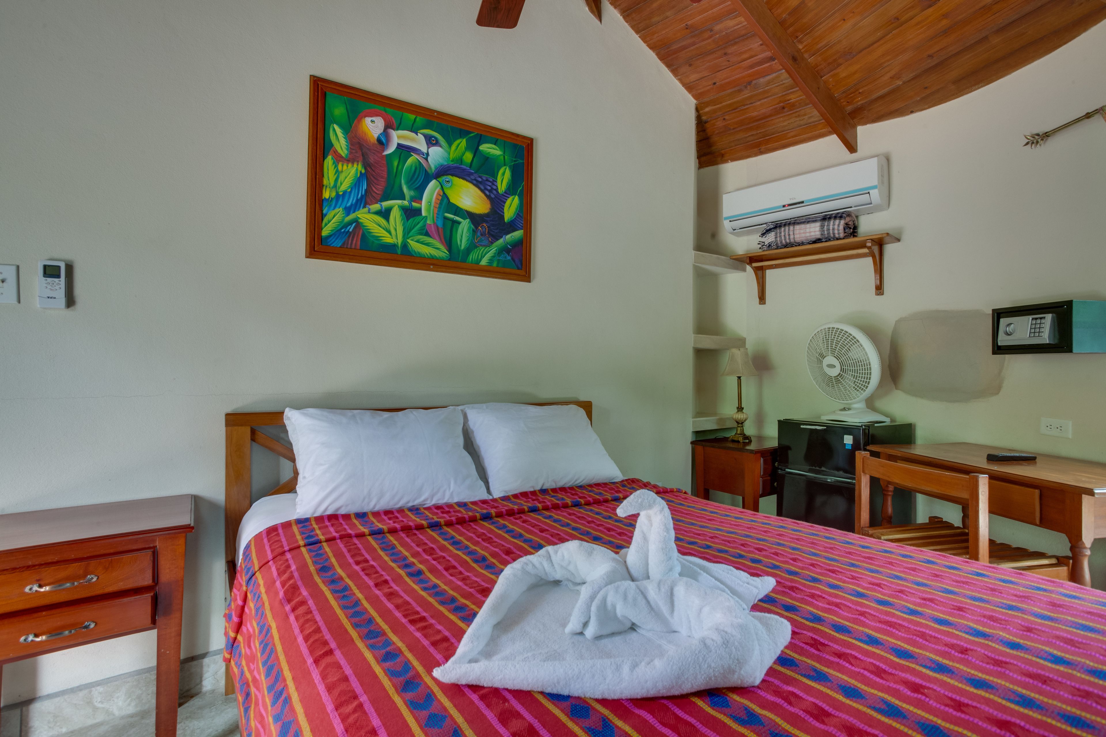 Habitación de hotel con cama, escritorio, silla, ventilador, aire acondicionado y un cuadro en la pared.