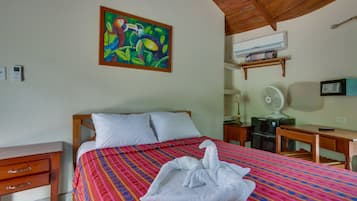 Habitación de hotel con cama, escritorio, silla, ventilador, aire acondicionado y un cuadro en la pared.