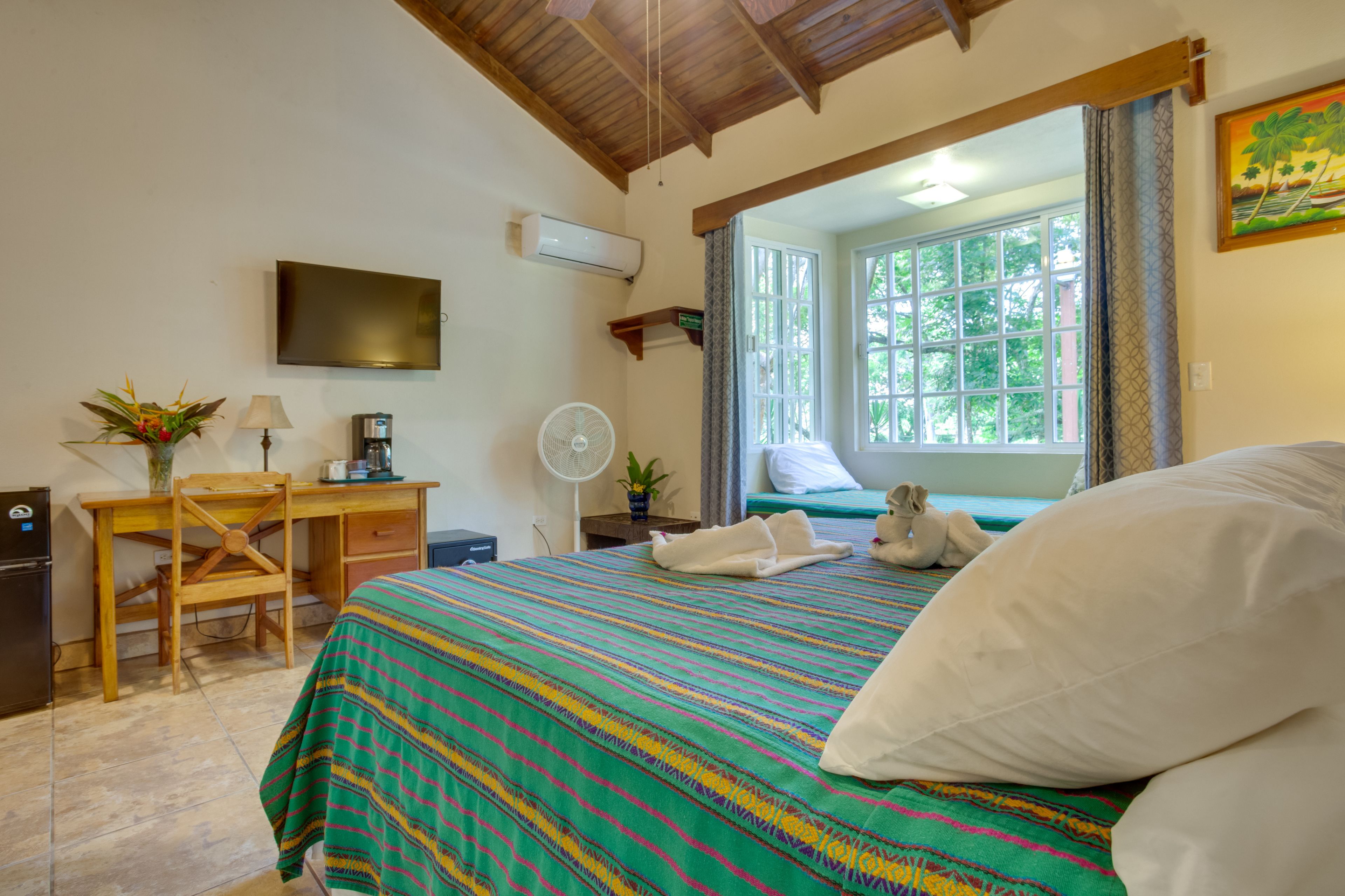 Habitación de hotel con cama, escritorio, silla, televisión y ventana con vista a árboles.