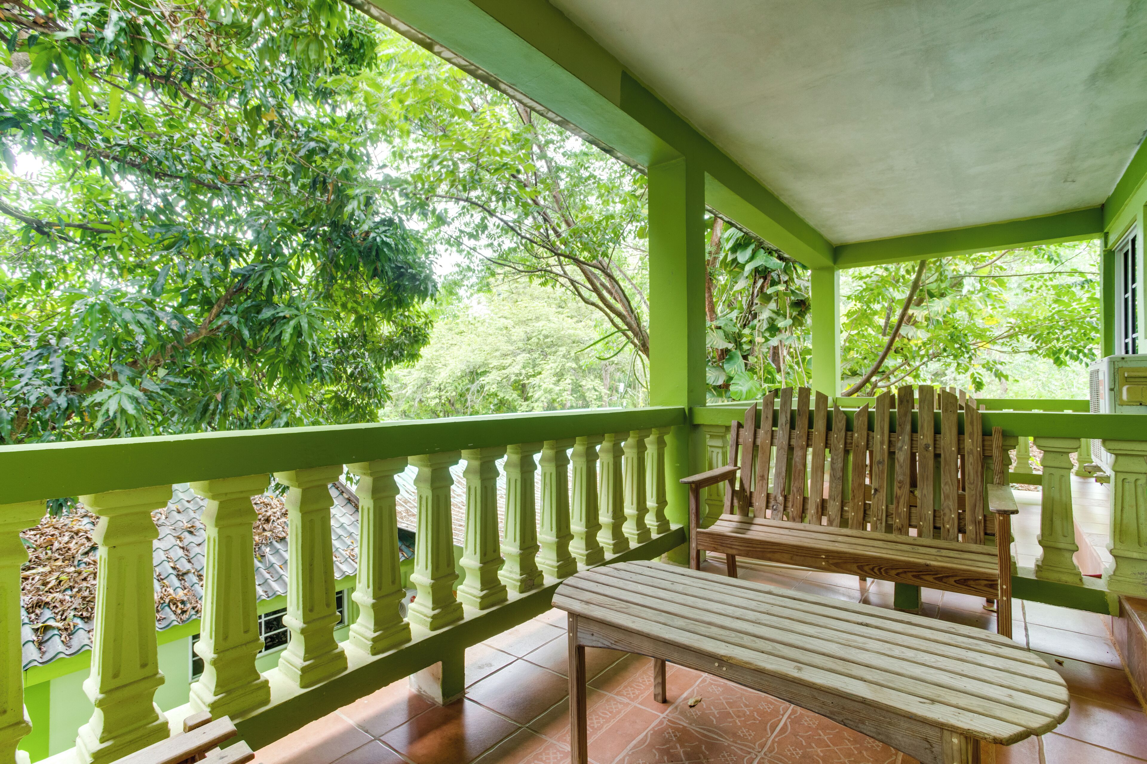 Un balcón con banca y mesa de madera, barandillas verdes y vistas de vegetación exuberante.