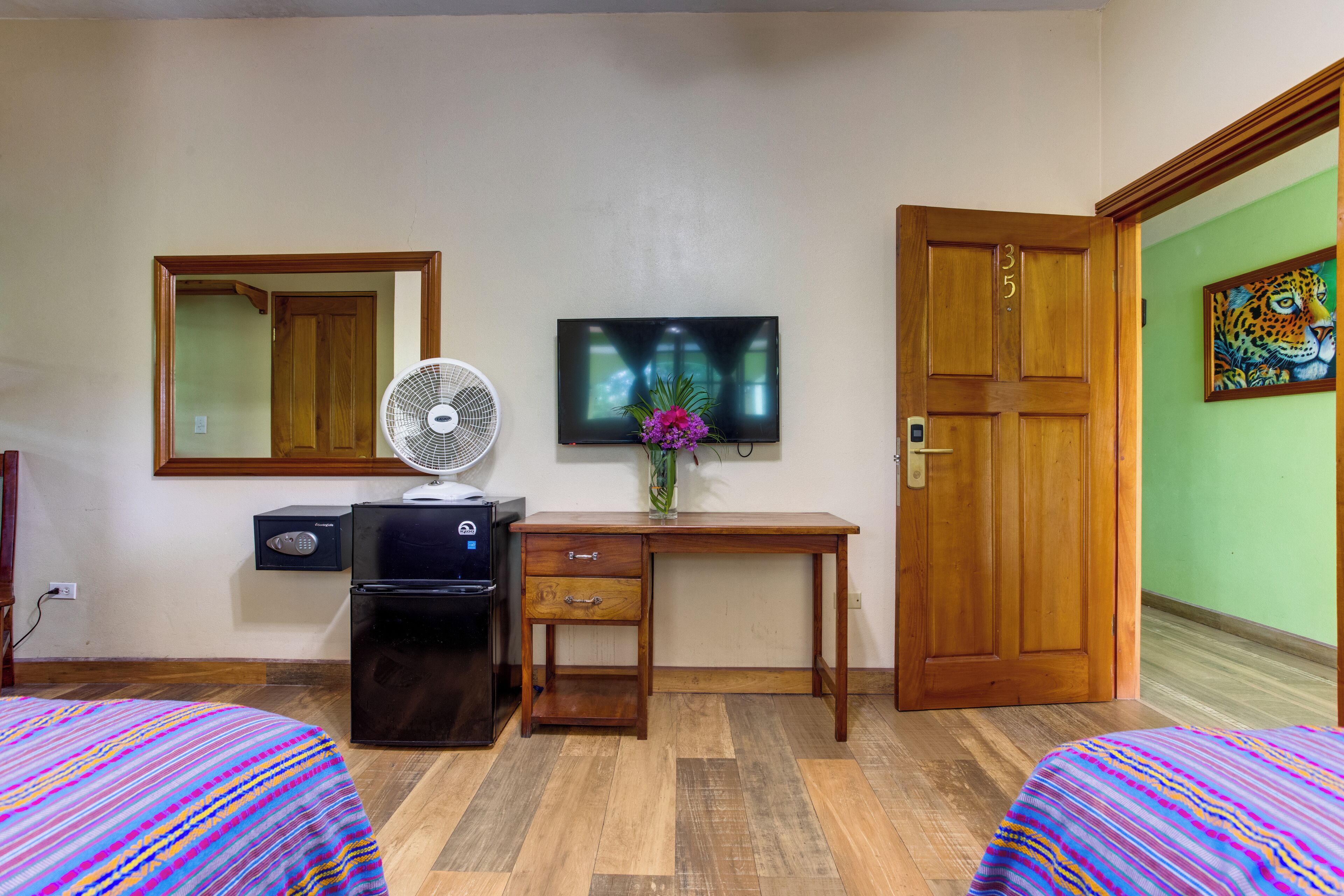 A hotel room with two beds, a desk with a TV, a fan, a refrigerator, and a door with a number.