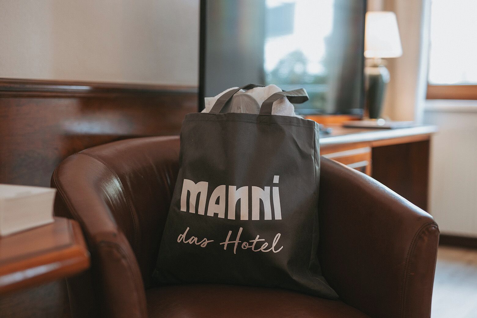 Un cojín gris oscuro con el texto "MANNI das Hotel" sobre un sillón de cuero marrón en una habitación de hotel.