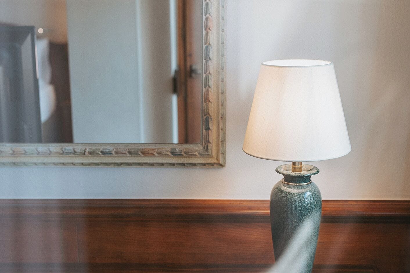 Eine Lampe mit weißem Lampenschirm auf einer Holzfläche, ein Spiegel mit dekorativem Rahmen und eine Holzvertäfelung darunter.