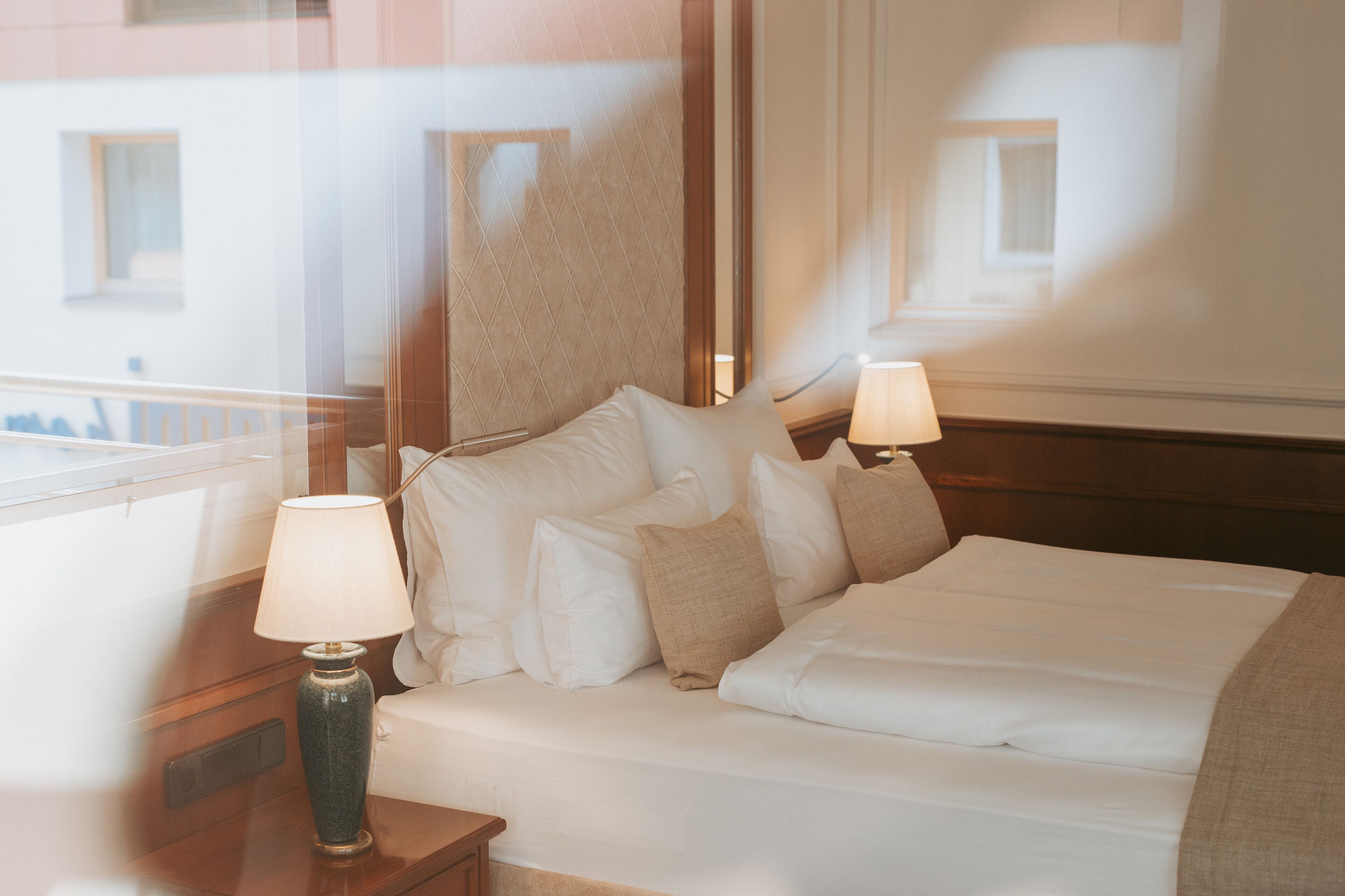 Una cama bien hecha con sábanas blancas y varios almohadones, dos lámparas de noche y vistas de un edificio a través de una ventana.