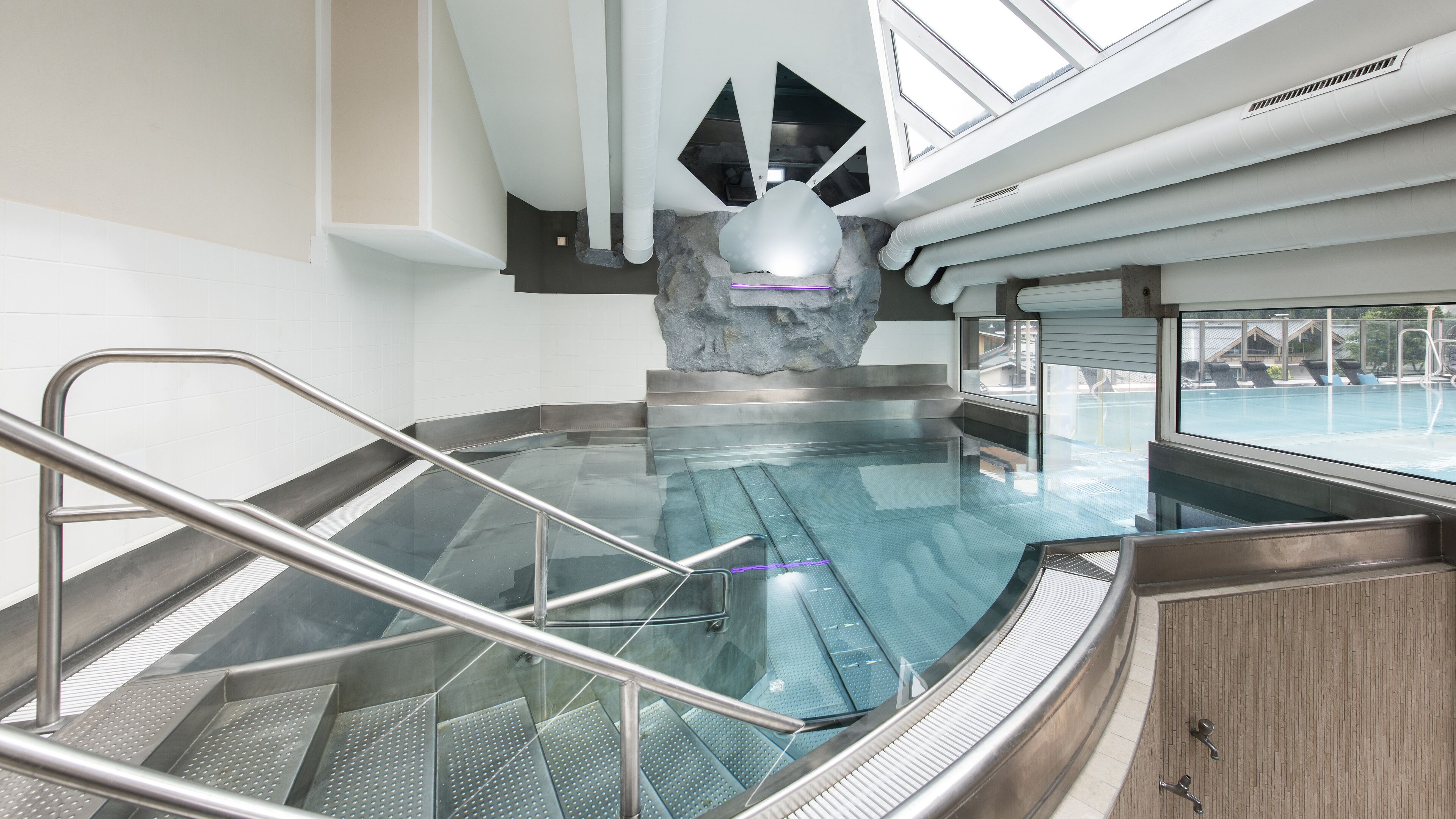 An indoor swimming pool with a glass barrier, a stone sculpture, and a view of a pool area through a window.