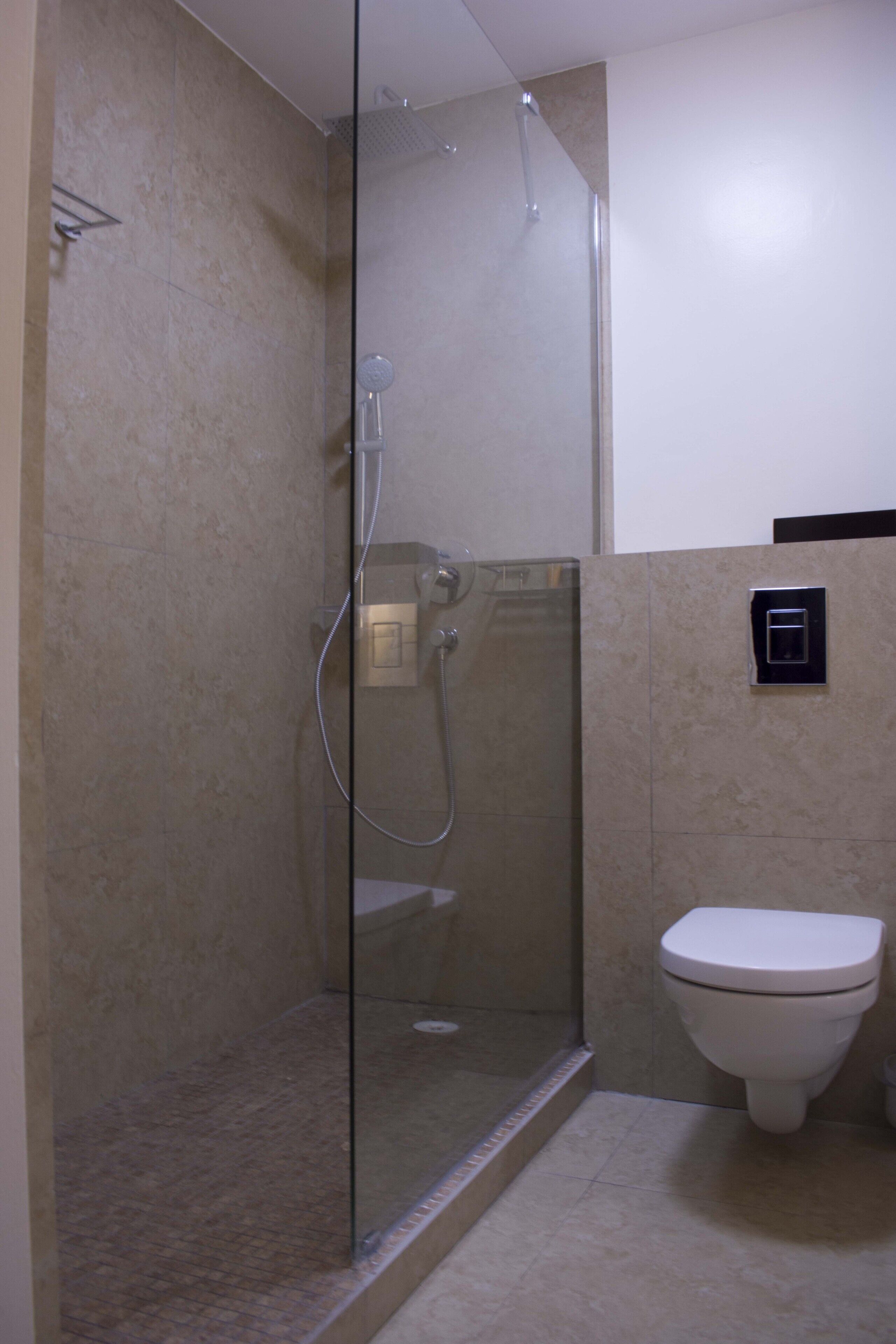 Et badeværelse med en glasafskærmet brusekabine, toilet og et panel til betjening af vand og varme monteret på væggen.