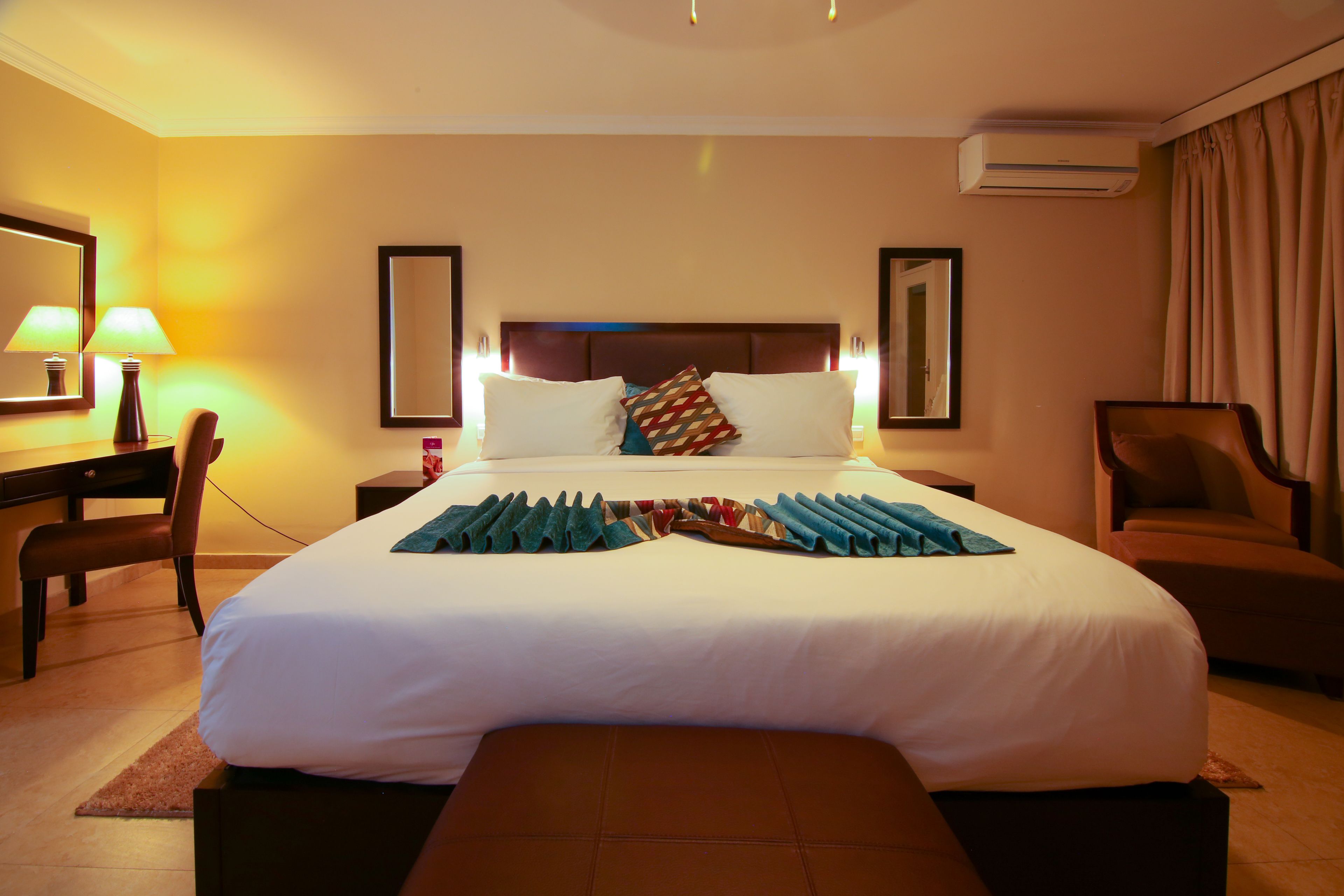 A hotel room with a large bed, bedside tables, a desk, a chair, and an air conditioning unit.