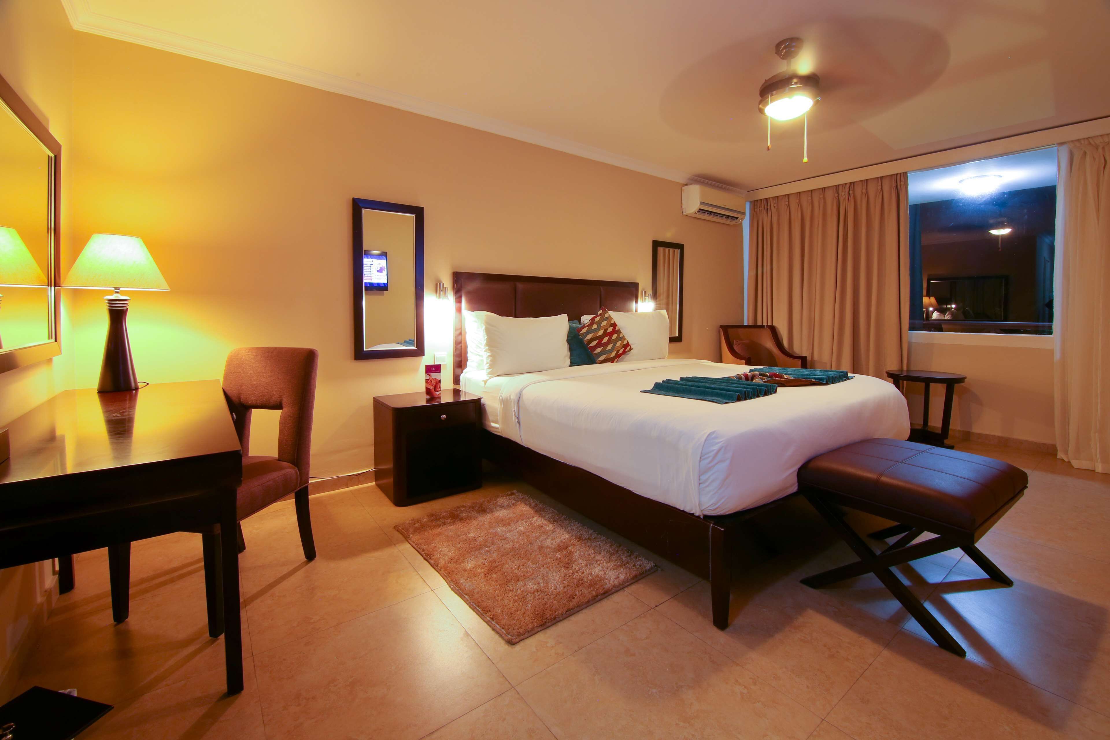 A hotel room with a bed, a desk, a chair, a bedside table, a lamp, a window with curtains, and a ceiling fan.