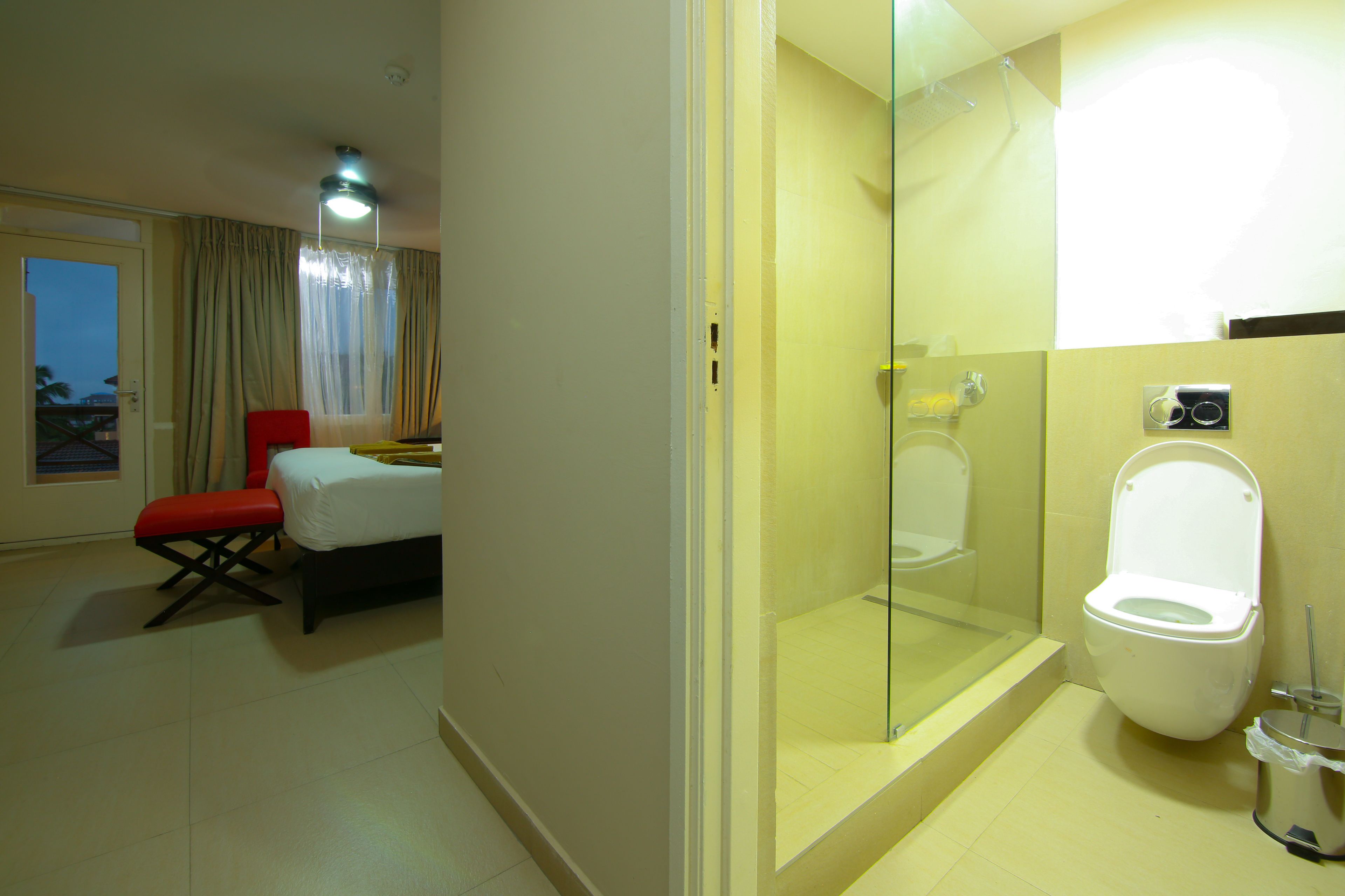 Habitación de hotel con cama, una silla roja, ducha con cerramiento de vidrio y un inodoro.