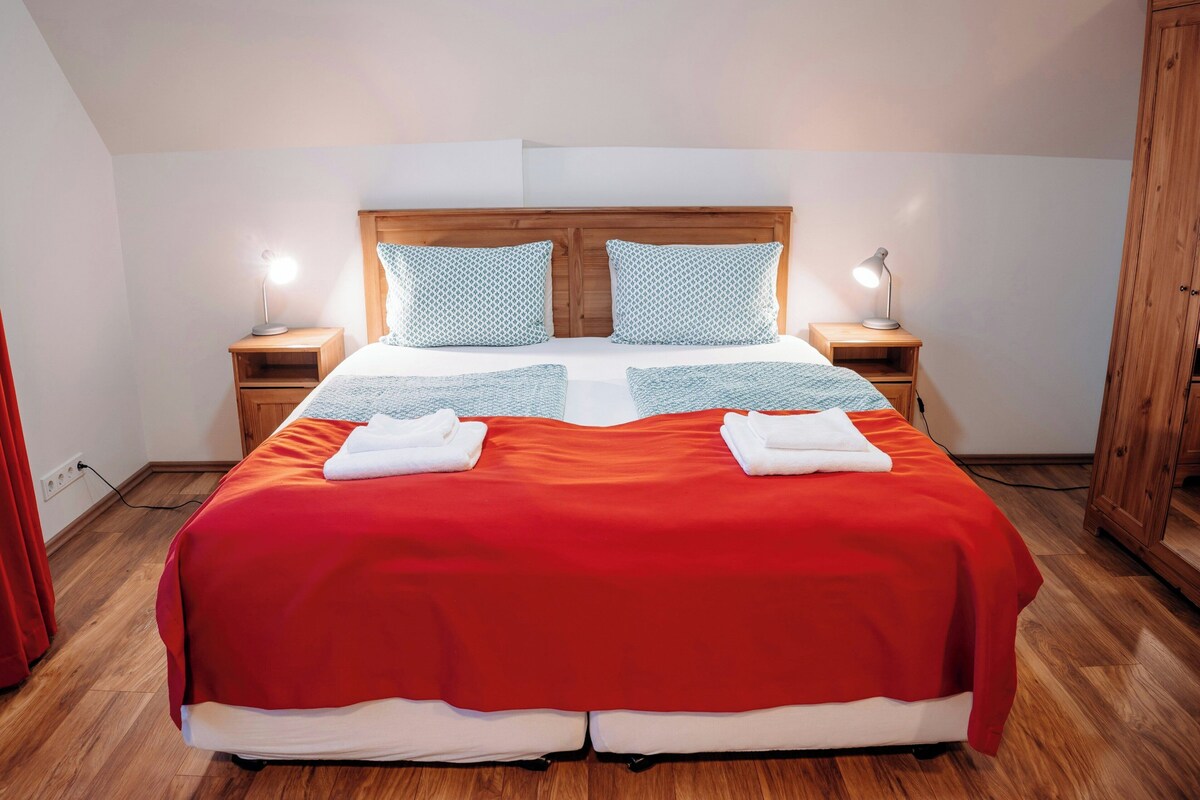 A neatly made bed with a red bedspread and two pillows, flanked by two bedside tables with lamps, in a room with wooden flooring and a wooden headboard.