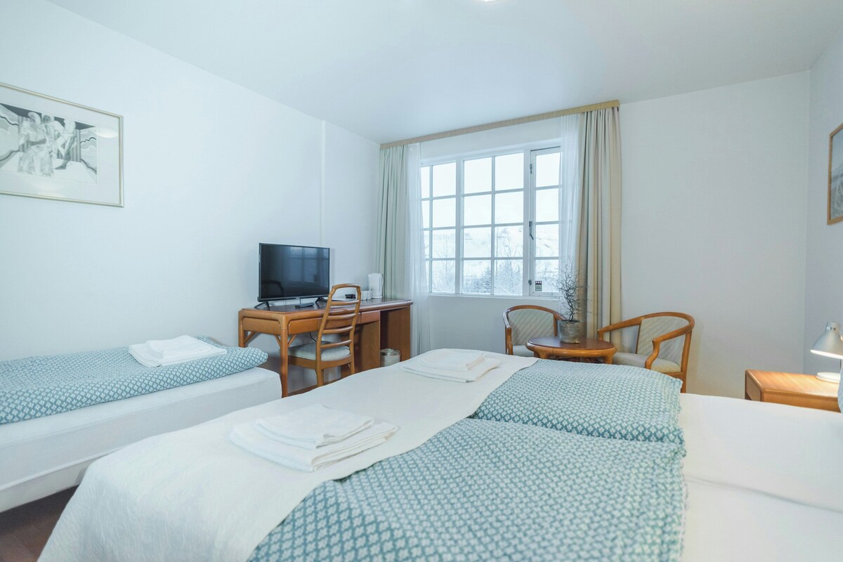 A hotel room with two single beds, a desk with a computer, a chair, a small table with a lamp, and a window with curtains.