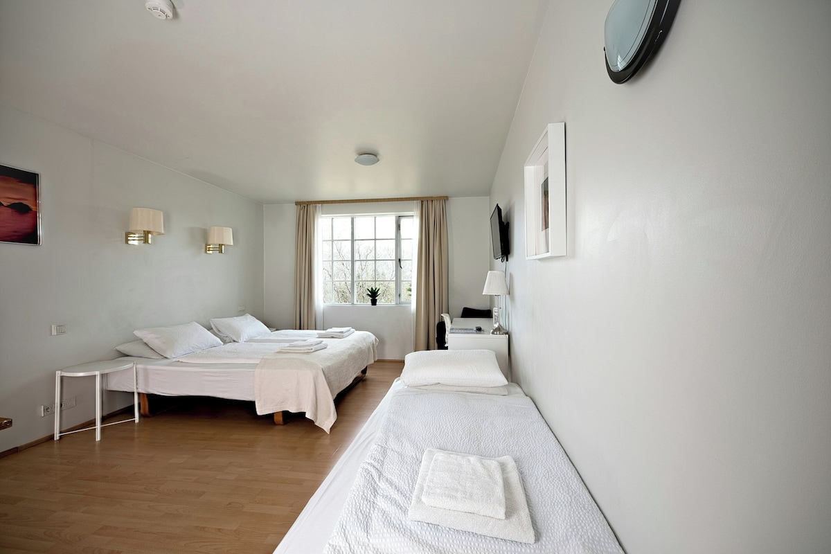 Habitación de hotel con dos camas, una ventana con cortinas, un escritorio con lámpara y un televisor montado en la pared.