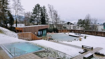 Un área de piscina moderna al aire libre con cubierta de madera, un pequeño edificio con puertas de cristal y un paisaje nevado.