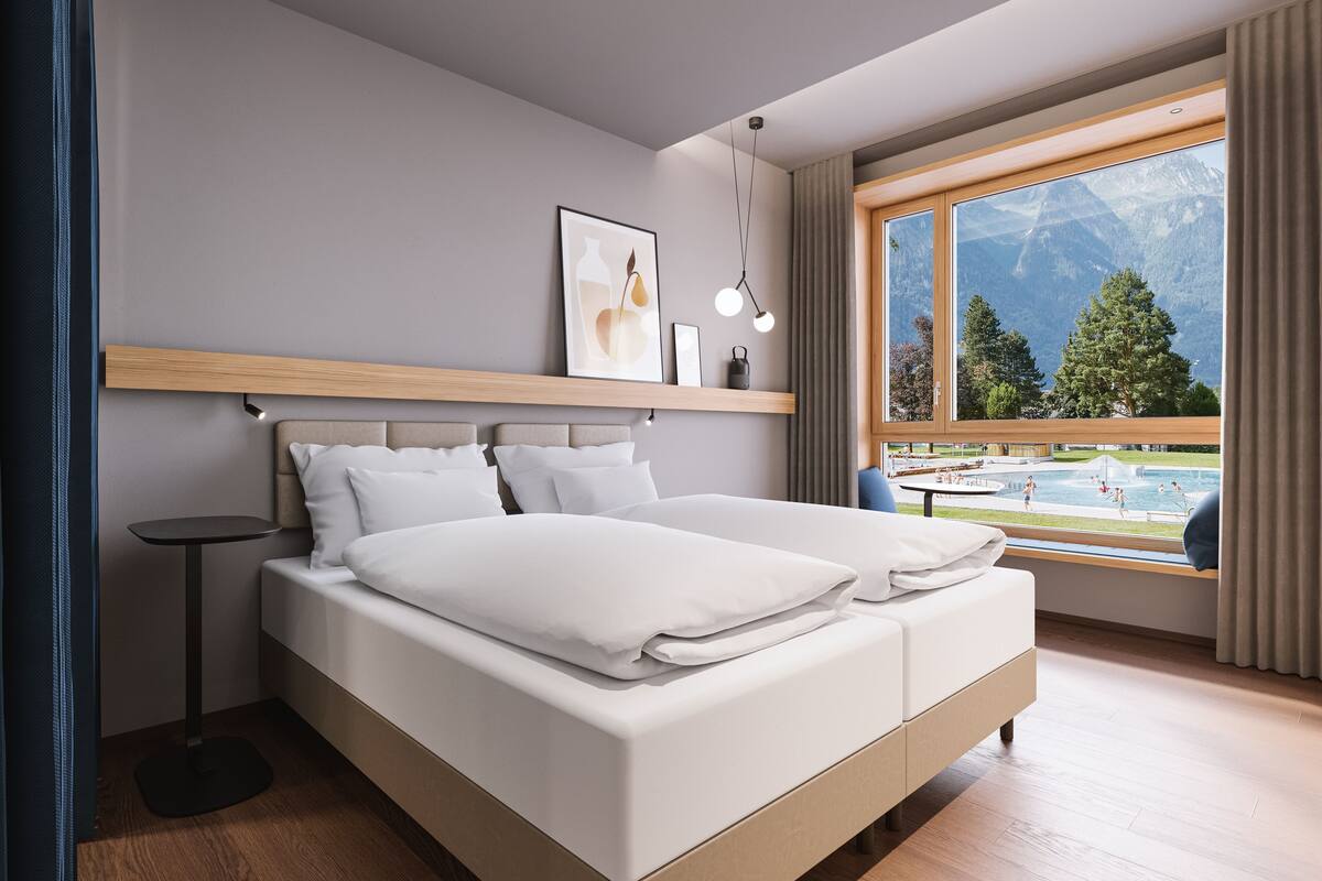 A modern hotel room with a large bed, a small side table, a window with a mountain view, and a wooden shelf above the bed.