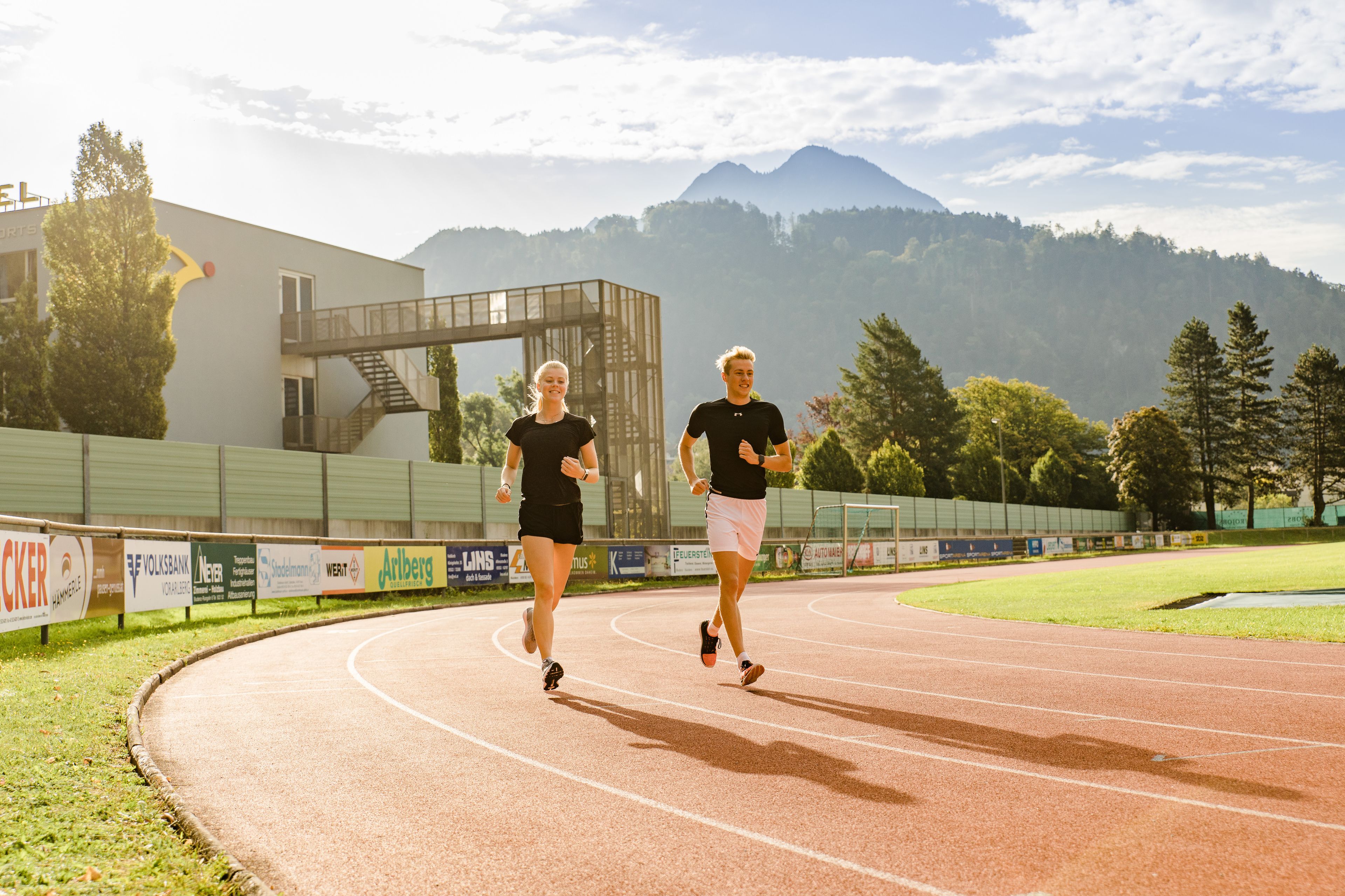 Pista de atletismo con dos corredores, rodeada por una valla y carteles publicitarios, con montañas al fondo.