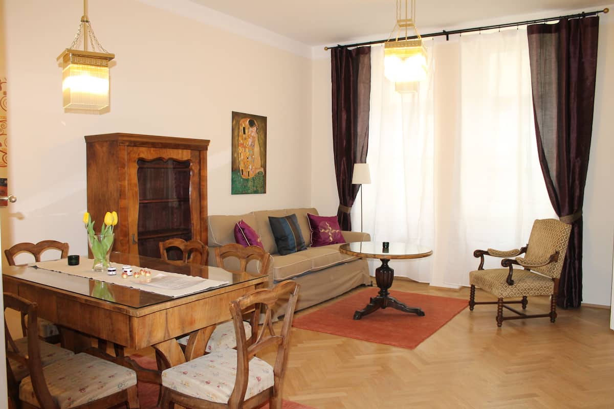 Uma sala de estar com uma mesa de jantar de madeira, cadeiras, um sofá, uma poltrona estampada e um quadro na parede.