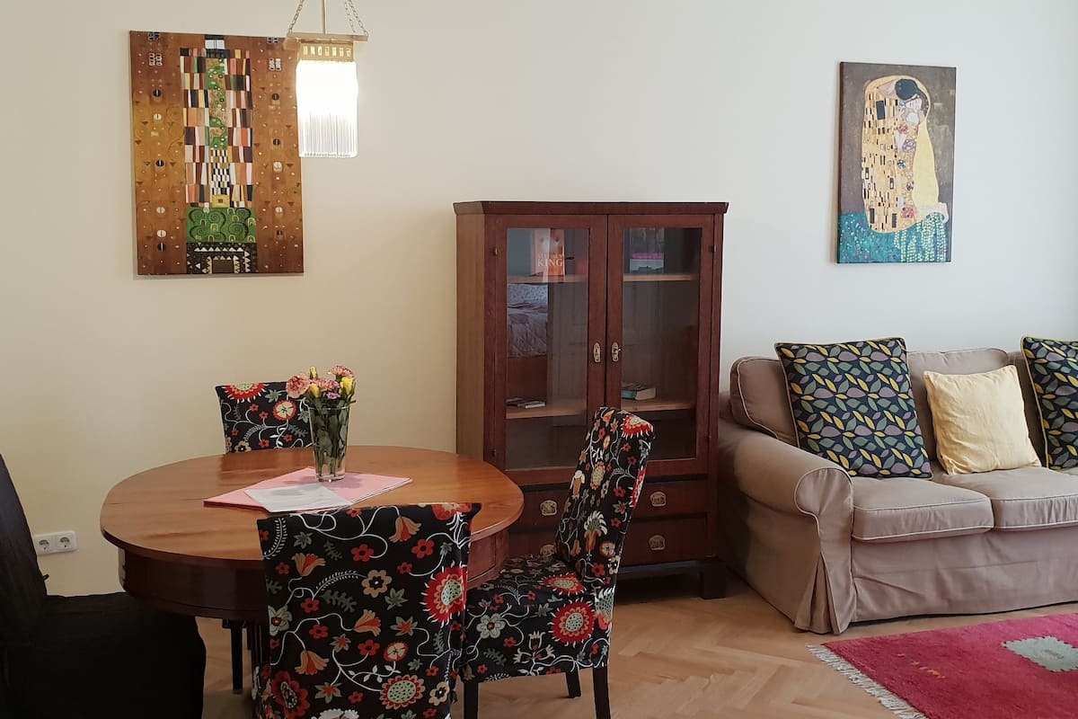 Uma sala de estar com uma mesa de jantar redonda, cadeiras estampadas com flores, um sofá com almofadas padronizadas, um armário e um quadro na parede.