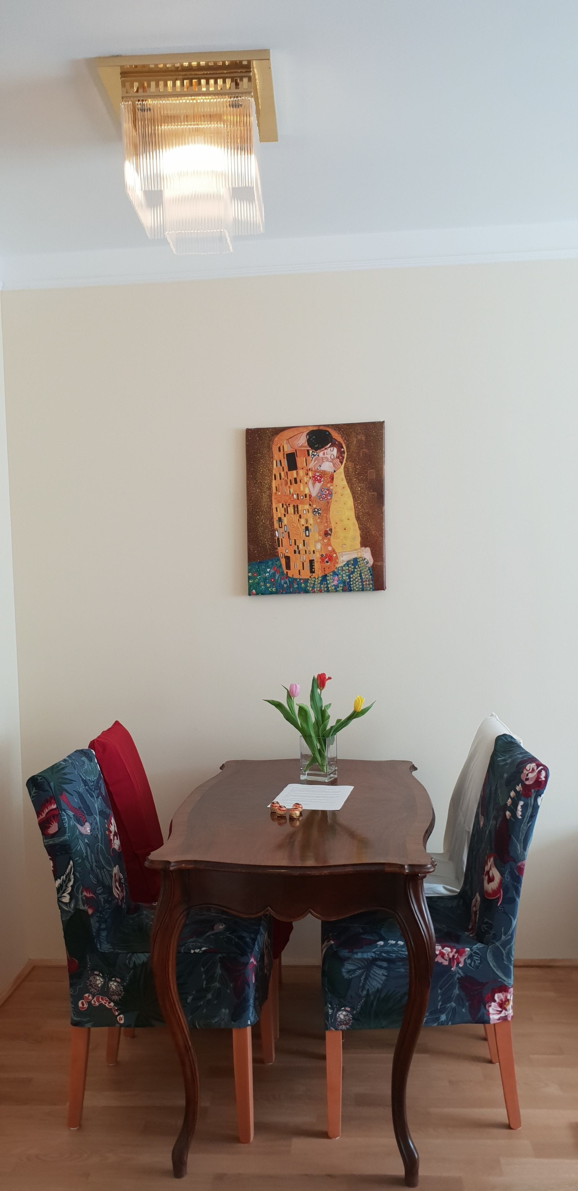 A dining area with a wooden table, floral chairs, a painting on the wall, and a ceiling light fixture.