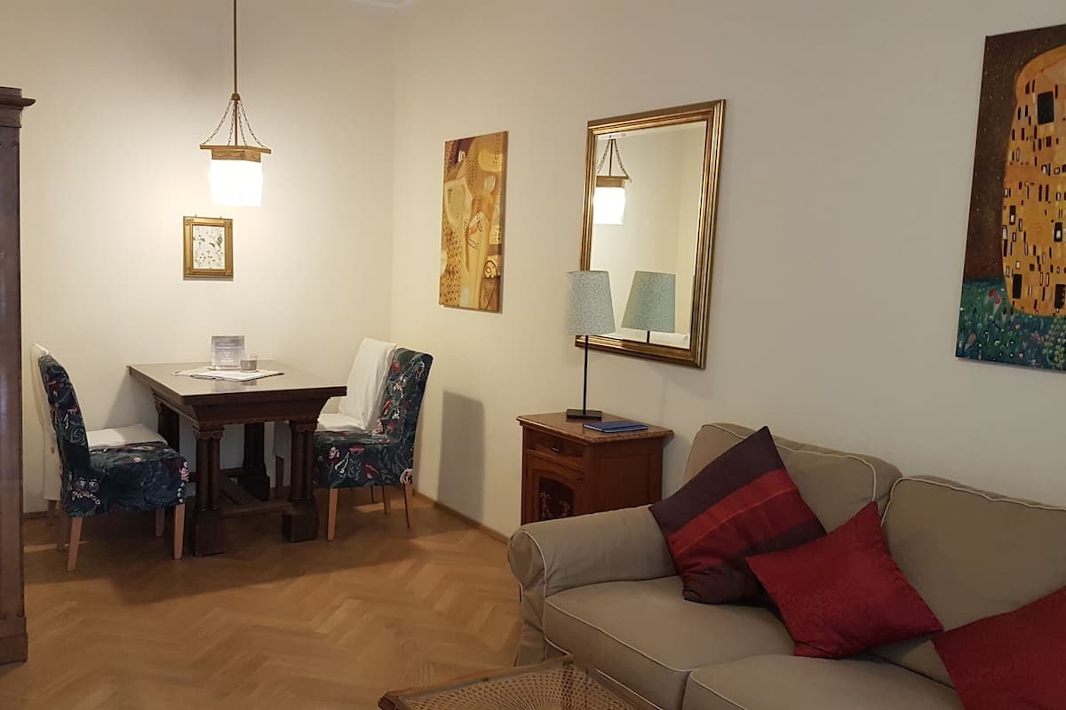Uma sala de estar com um armário de madeira, uma mesa de jantar com cadeiras, um sofá com almofadas, um abajur e um quadro na parede.