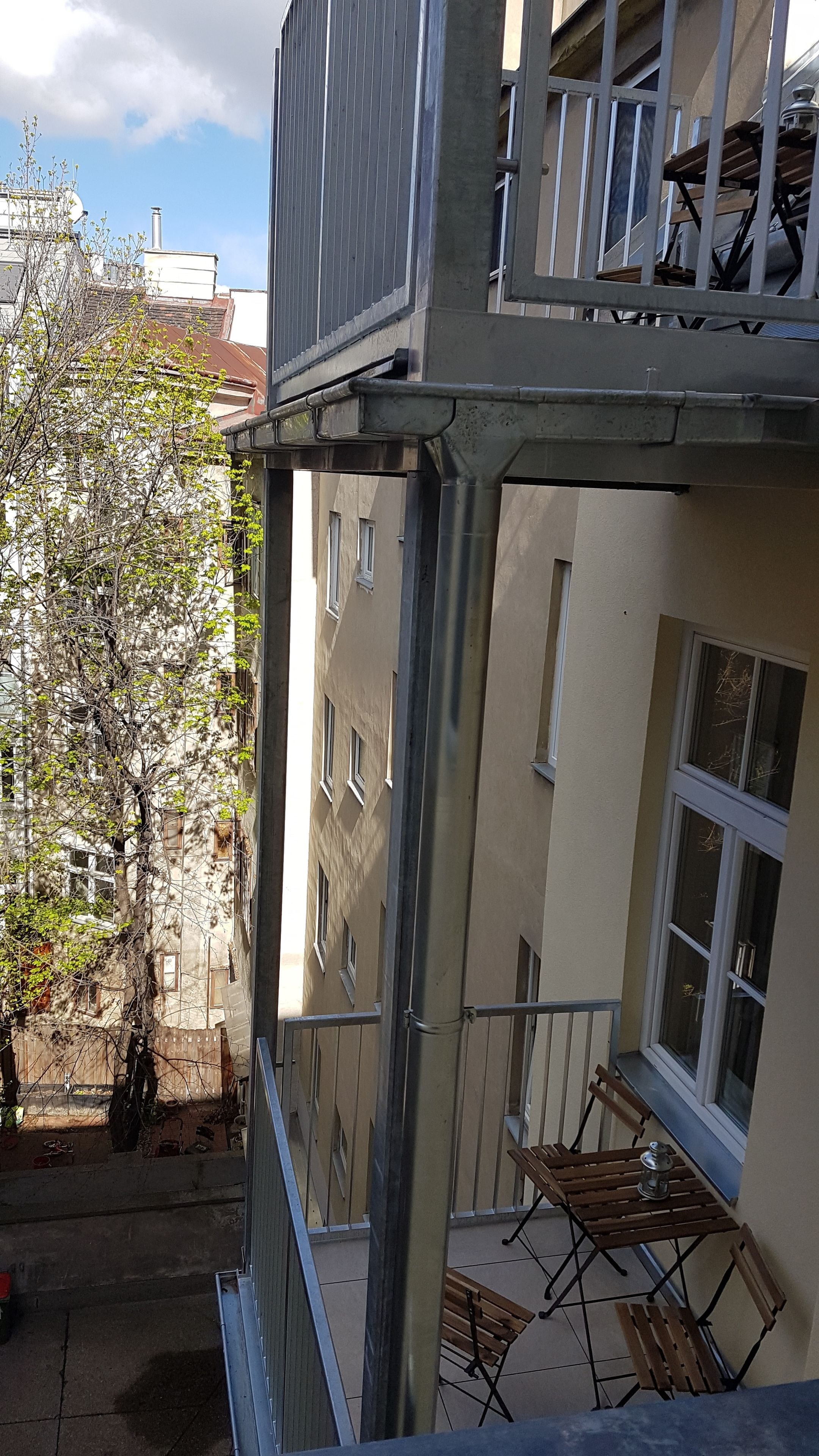 Ein mehrgeschossiges Gebäude mit Balkon, Außenbestuhlung und einem Baum.