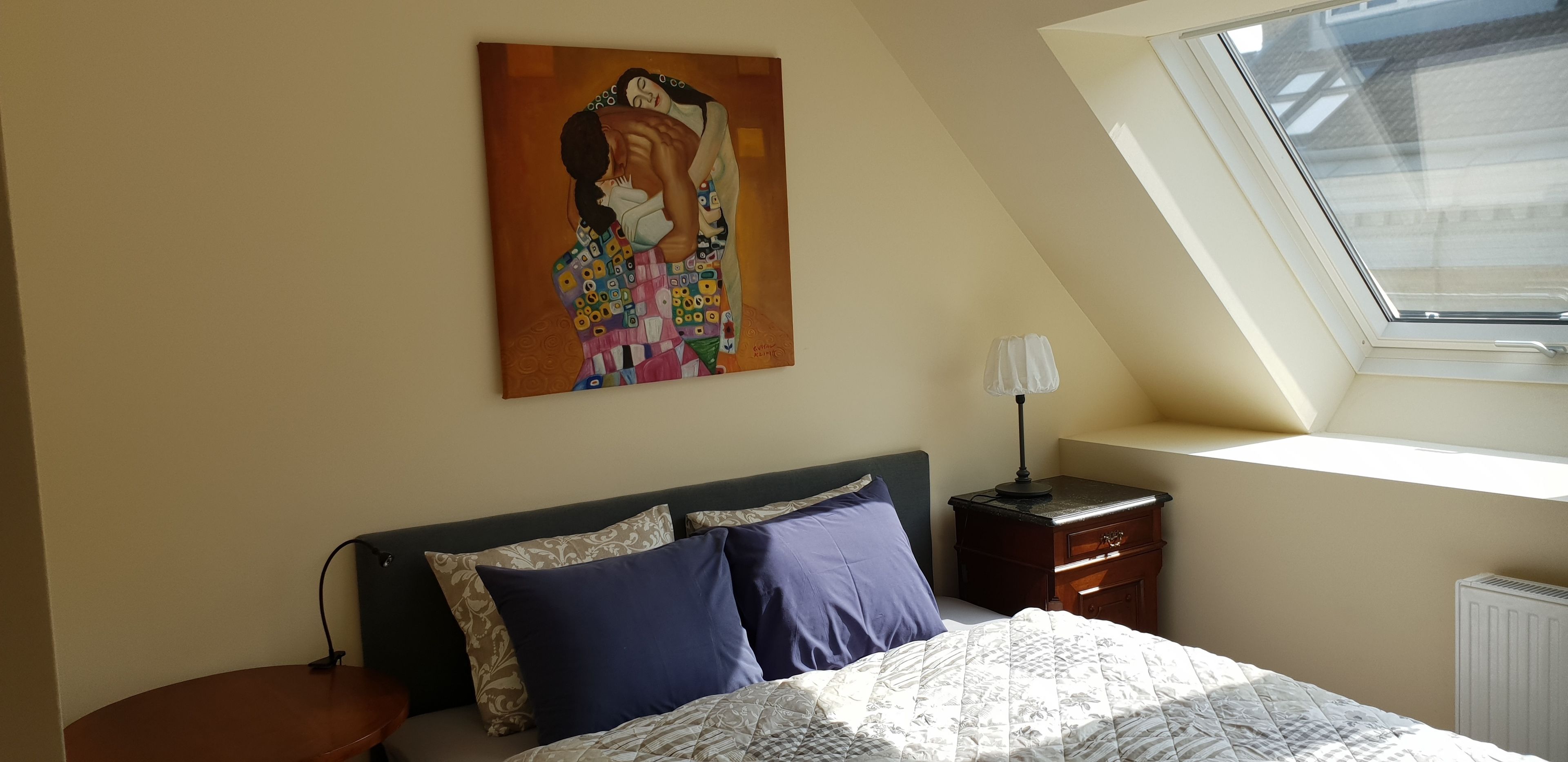 A bedroom with a bed, a nightstand, a lamp, a painting, and a skylight.